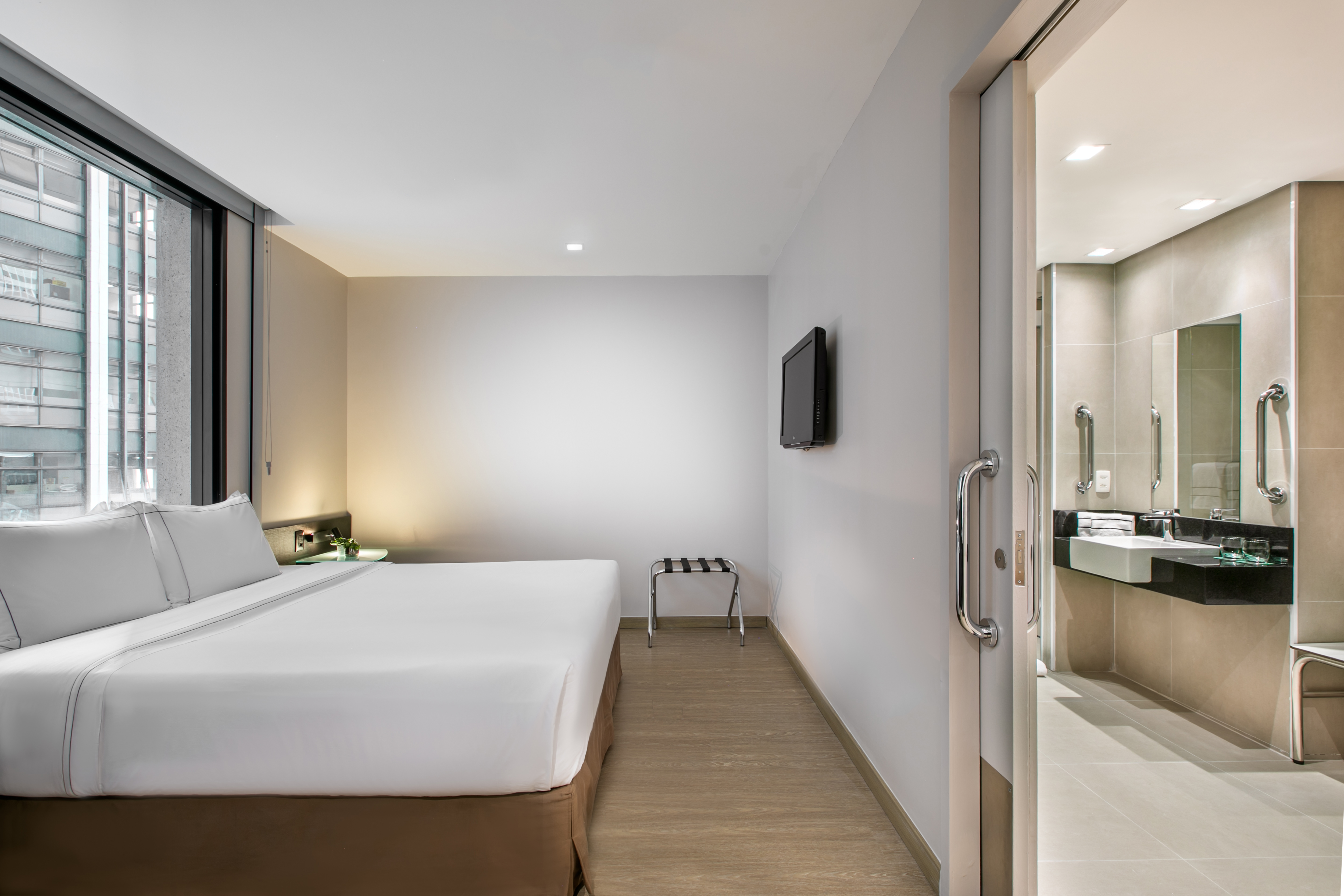 Accessible modern hotel bedroom with white bed, city view, and adjoining bathroom.