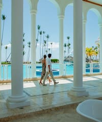 a man and woman walking on a walkway with columns and a pool