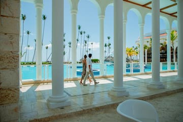 a man and woman walking on a walkway with columns and a pool