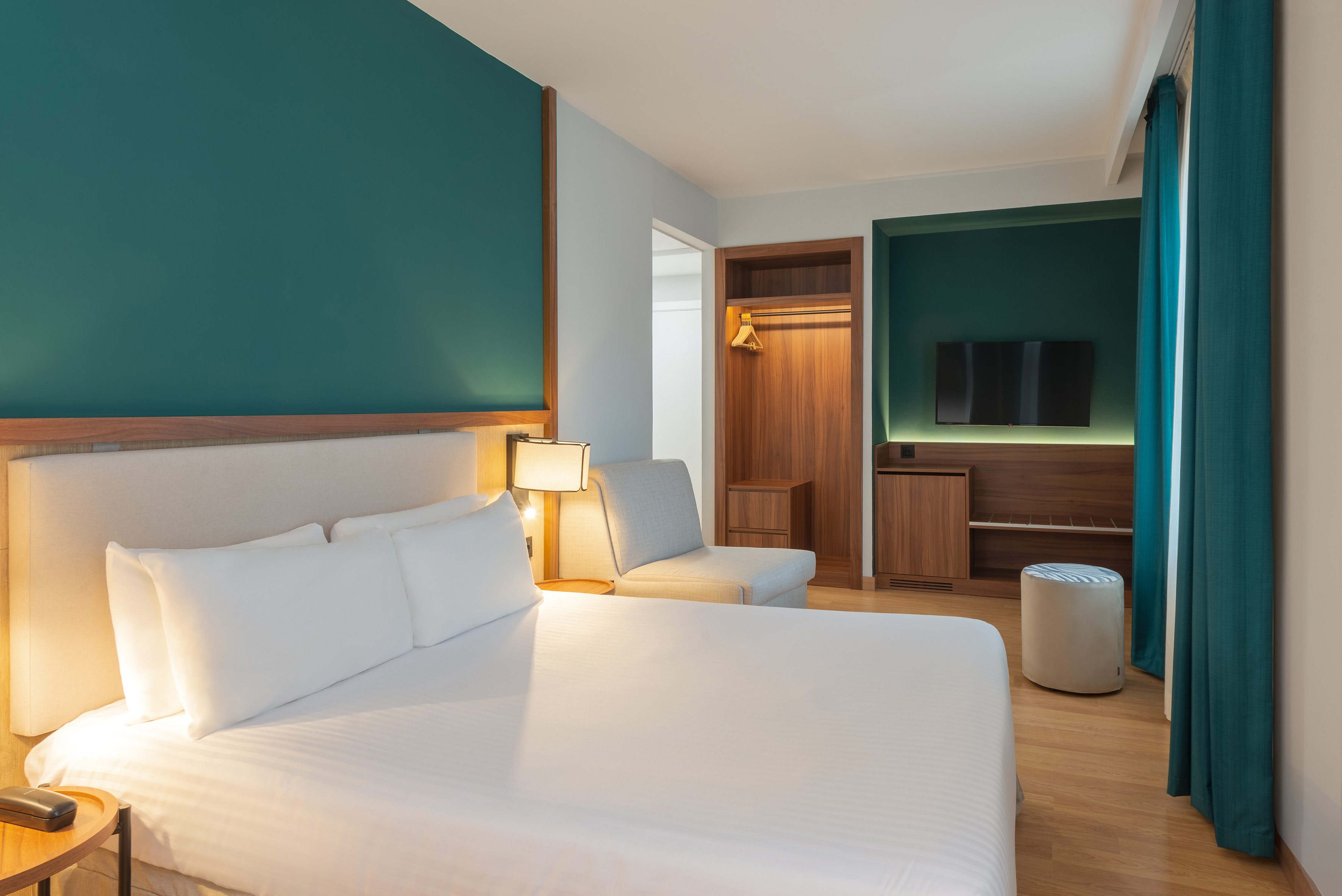 Modern hotel bedroom with bed, teal walls, wood flooring, and dark wood furniture.