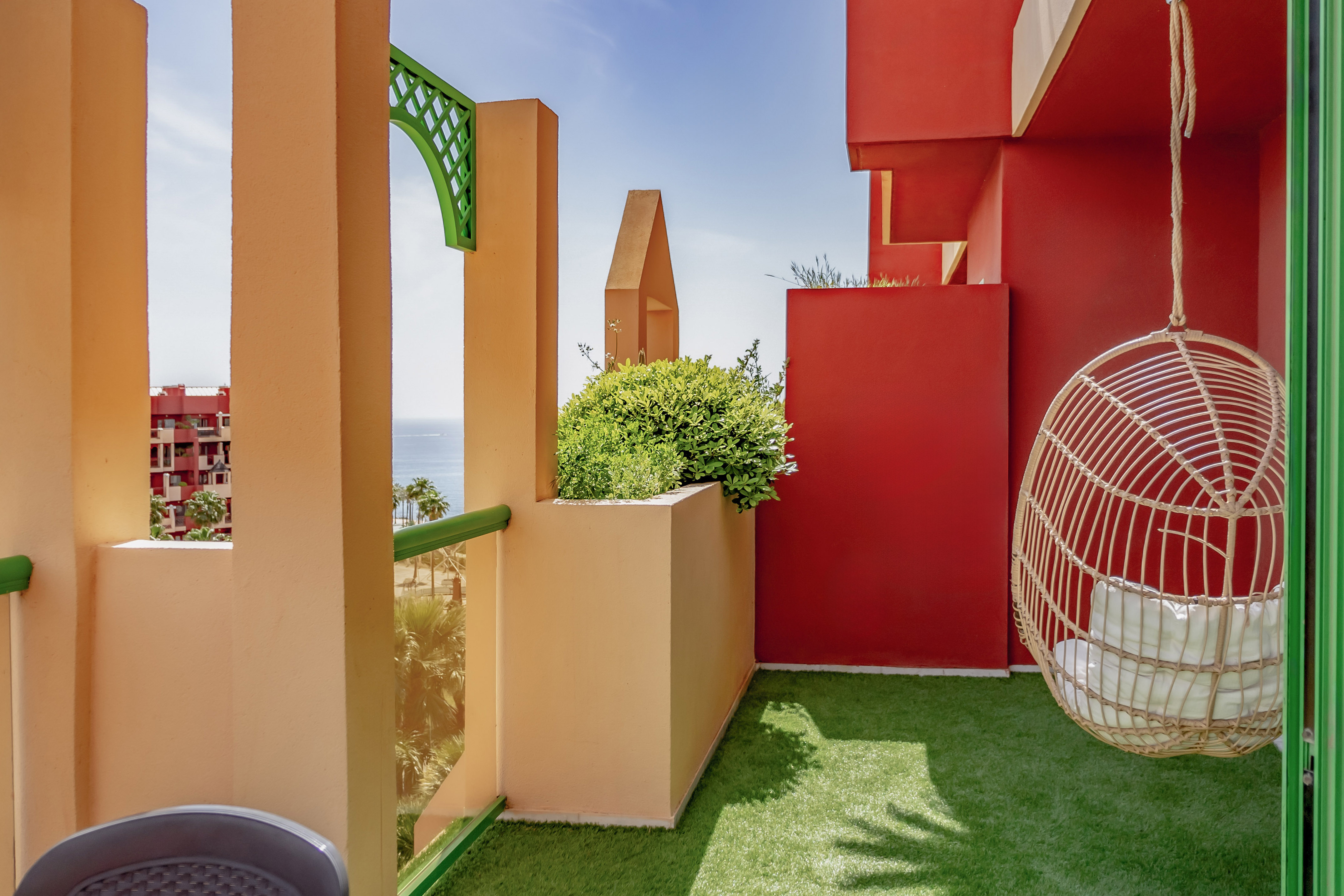 Vibrant balcony with red, peach walls, rattan hanging chair, ocean view, green turf.