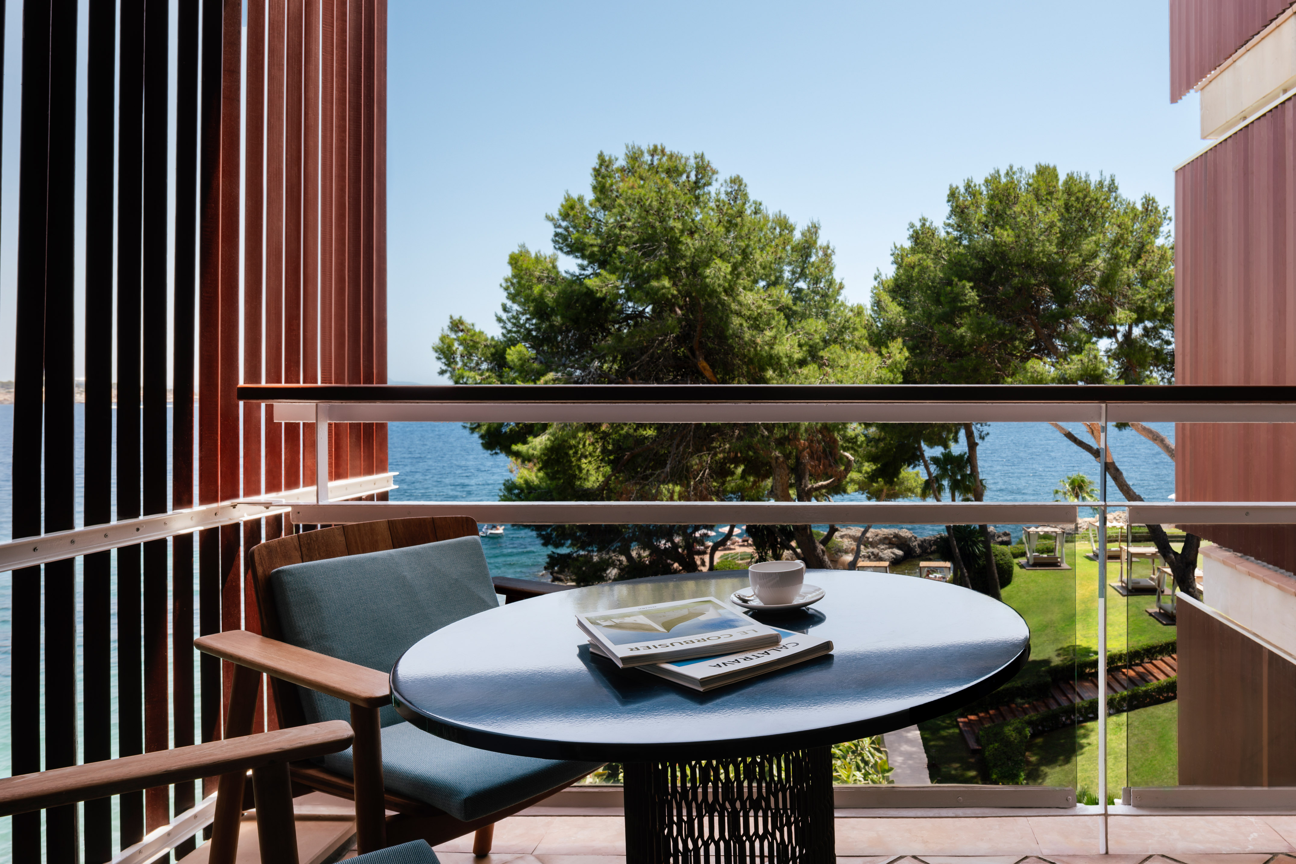 Balcony sea view with outdoor table, coffee, magazines, and lush pine trees.