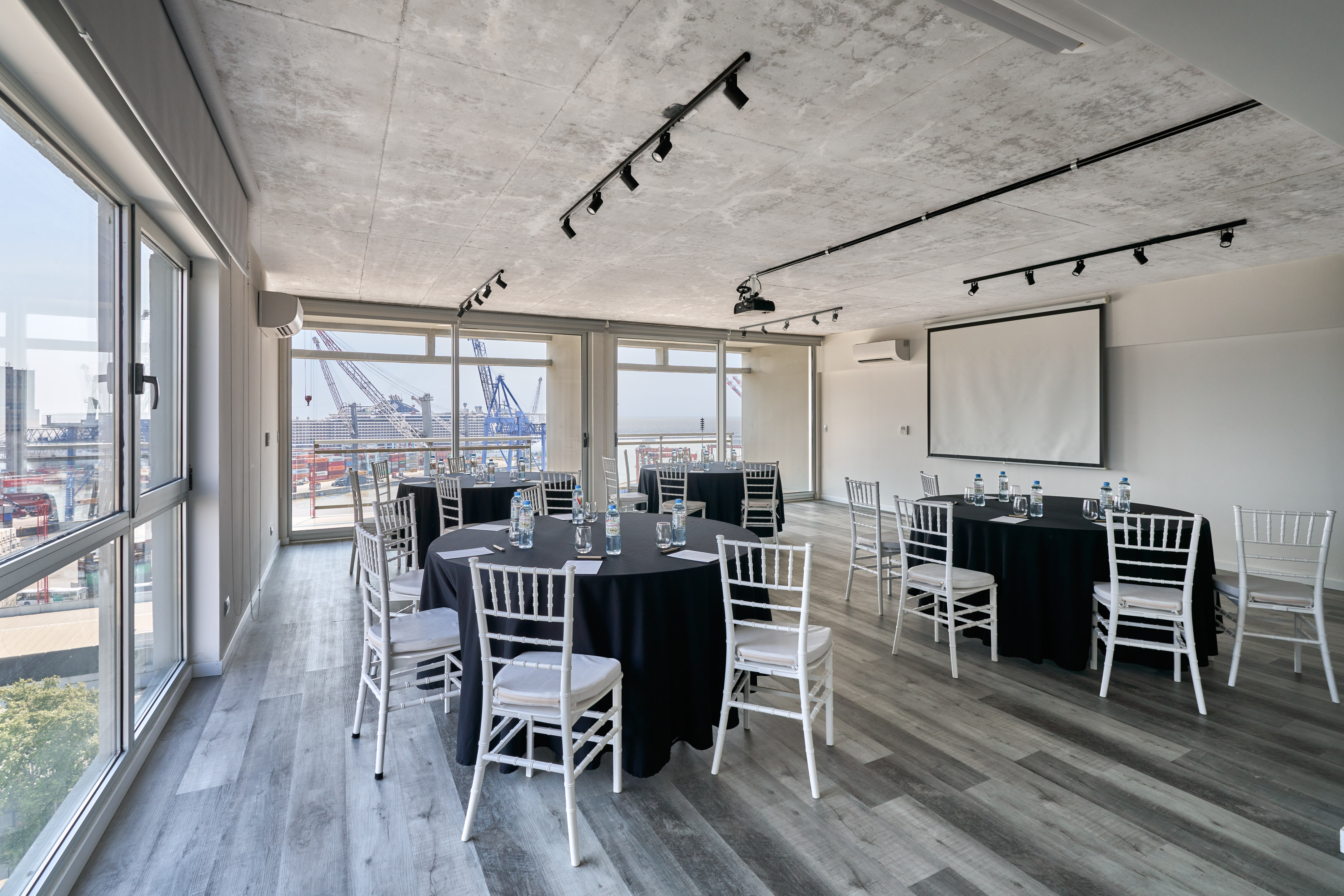 Modern meeting room with harbor view, round tables, white chairs, projector screen.