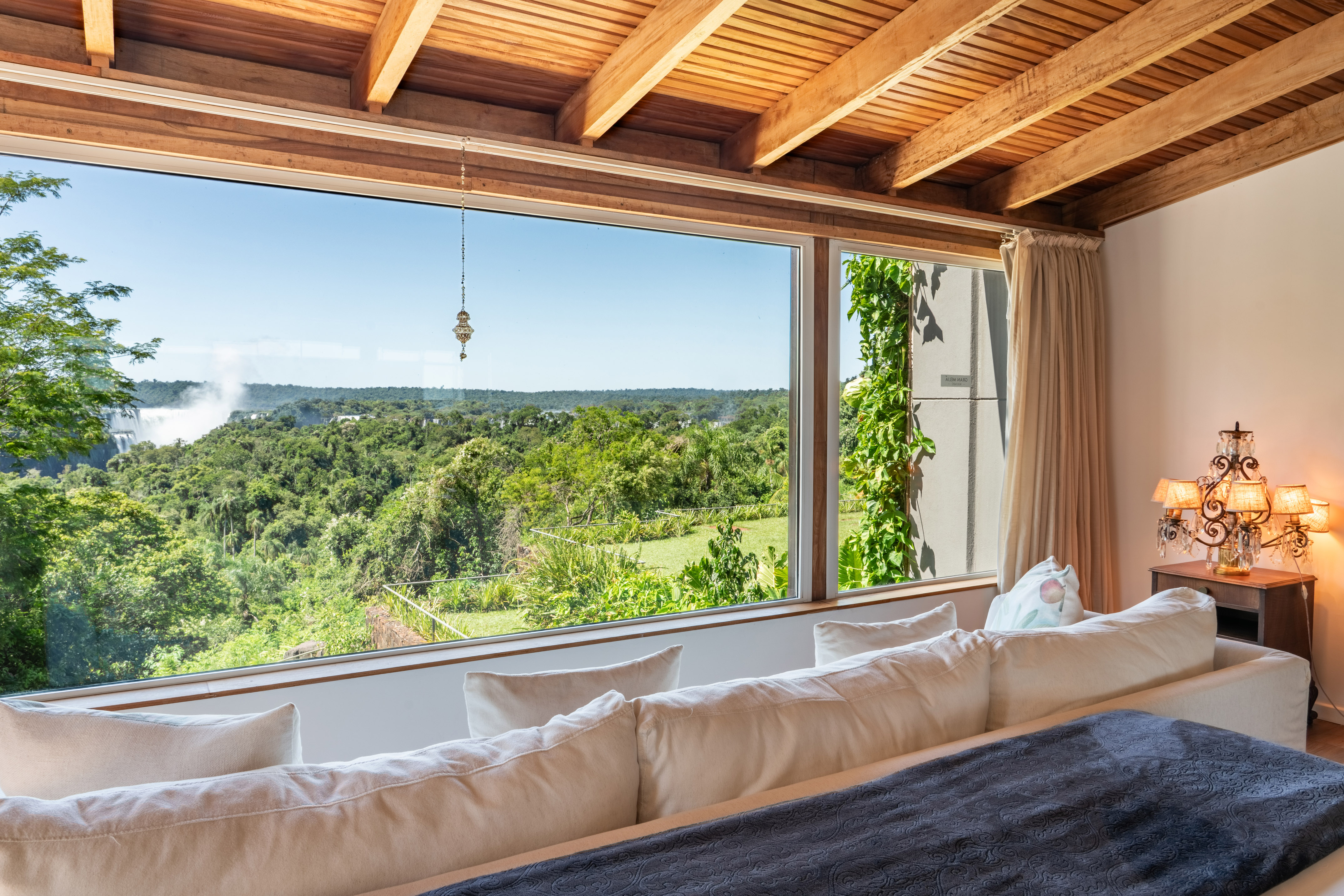 Panoramic bedroom view of distant waterfall, lush jungle, and wooden ceiling.