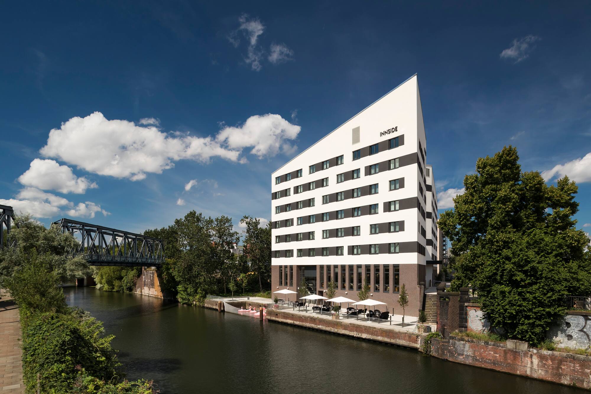 Angular INNSIDE hotel by river, outdoor patio, and metal bridge.