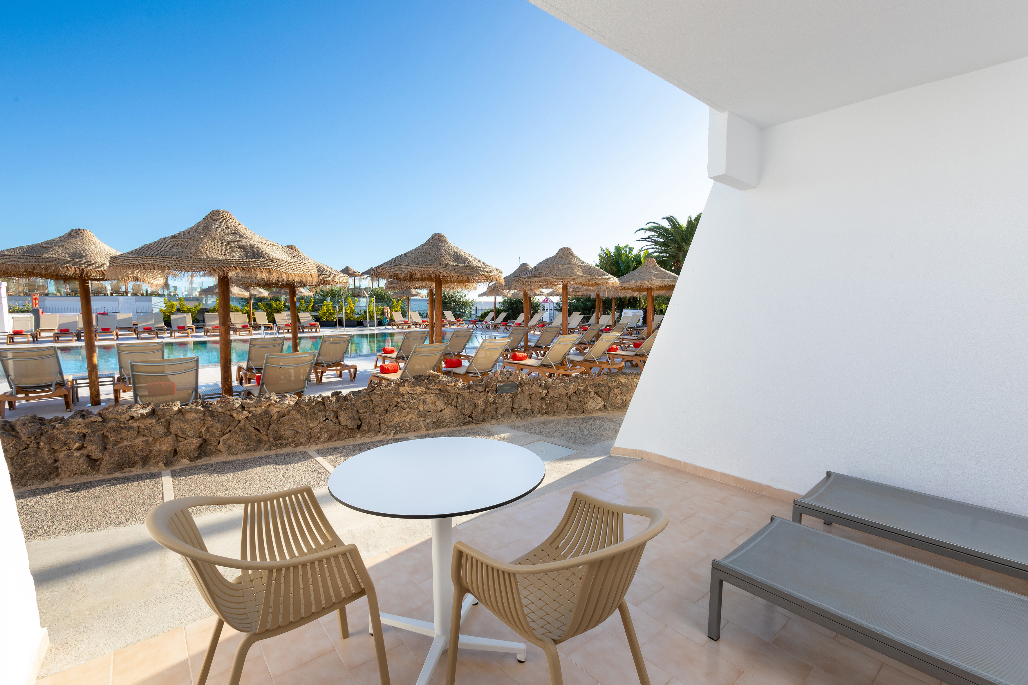 Sunny hotel terrace with pool view, sun loungers, and thatched umbrellas.