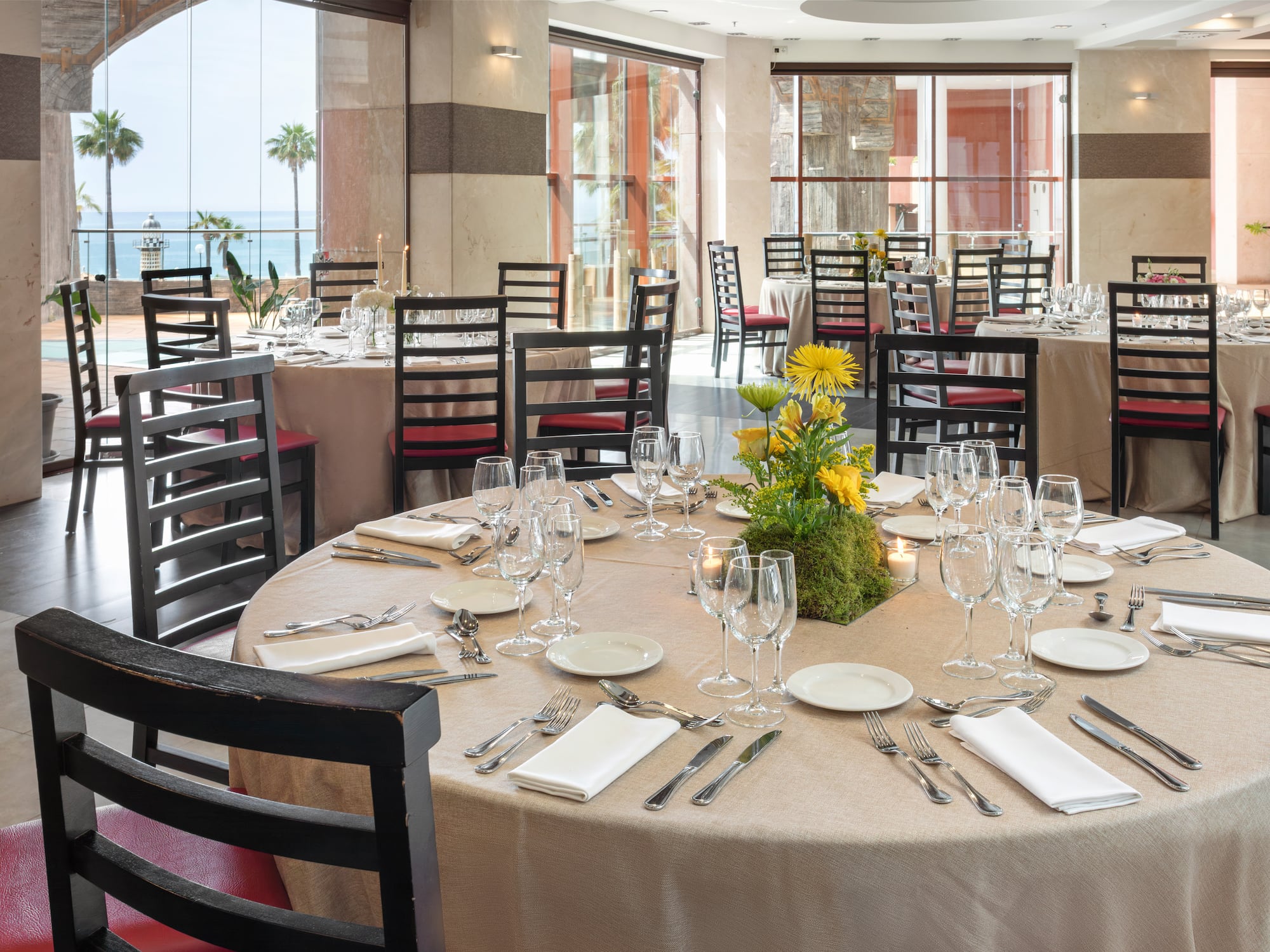 Upscale restaurant dining room with ocean view, set tables, and palm trees.