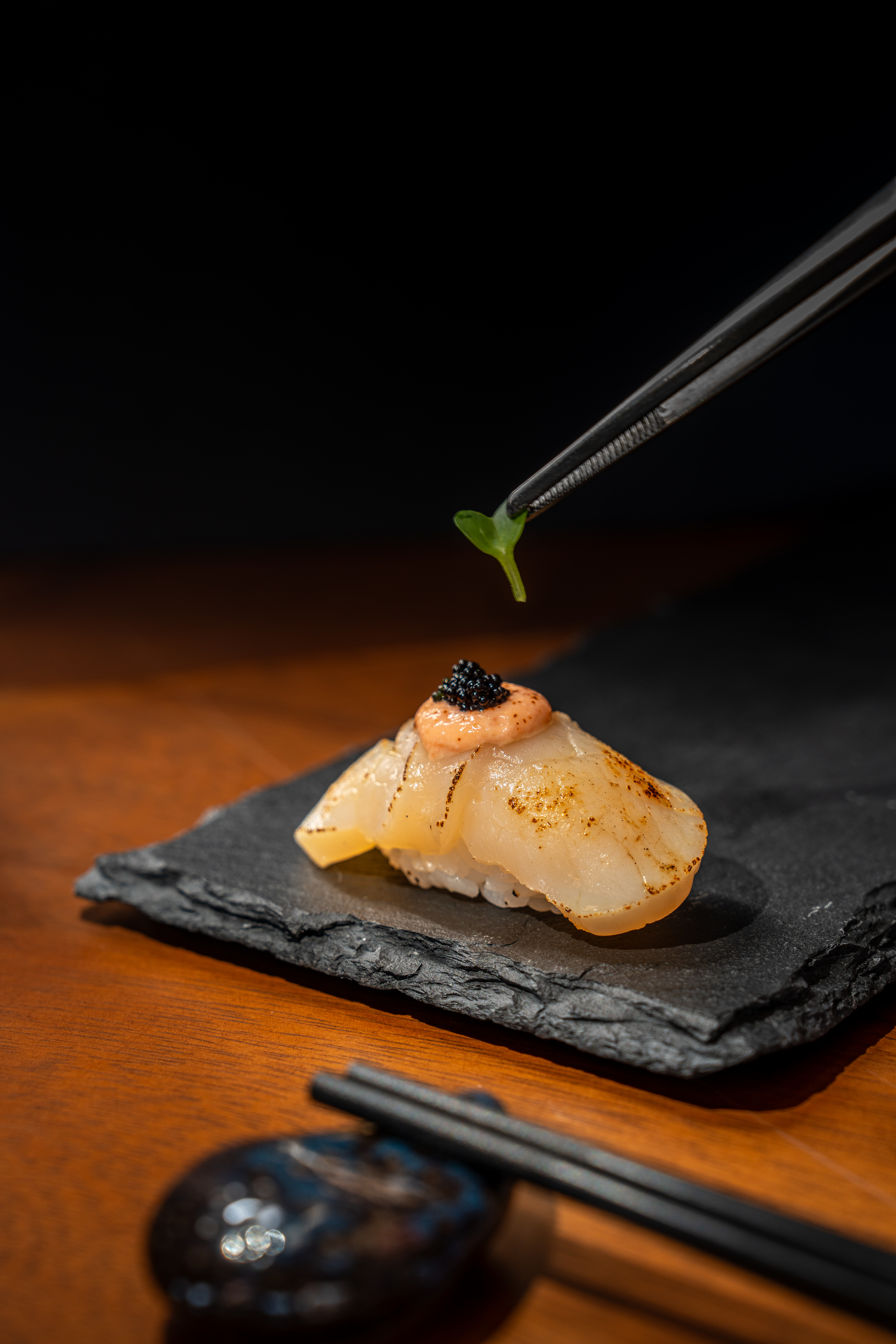 Gourmet seared scallop nigiri sushi with caviar, tweezers adding microgreen.