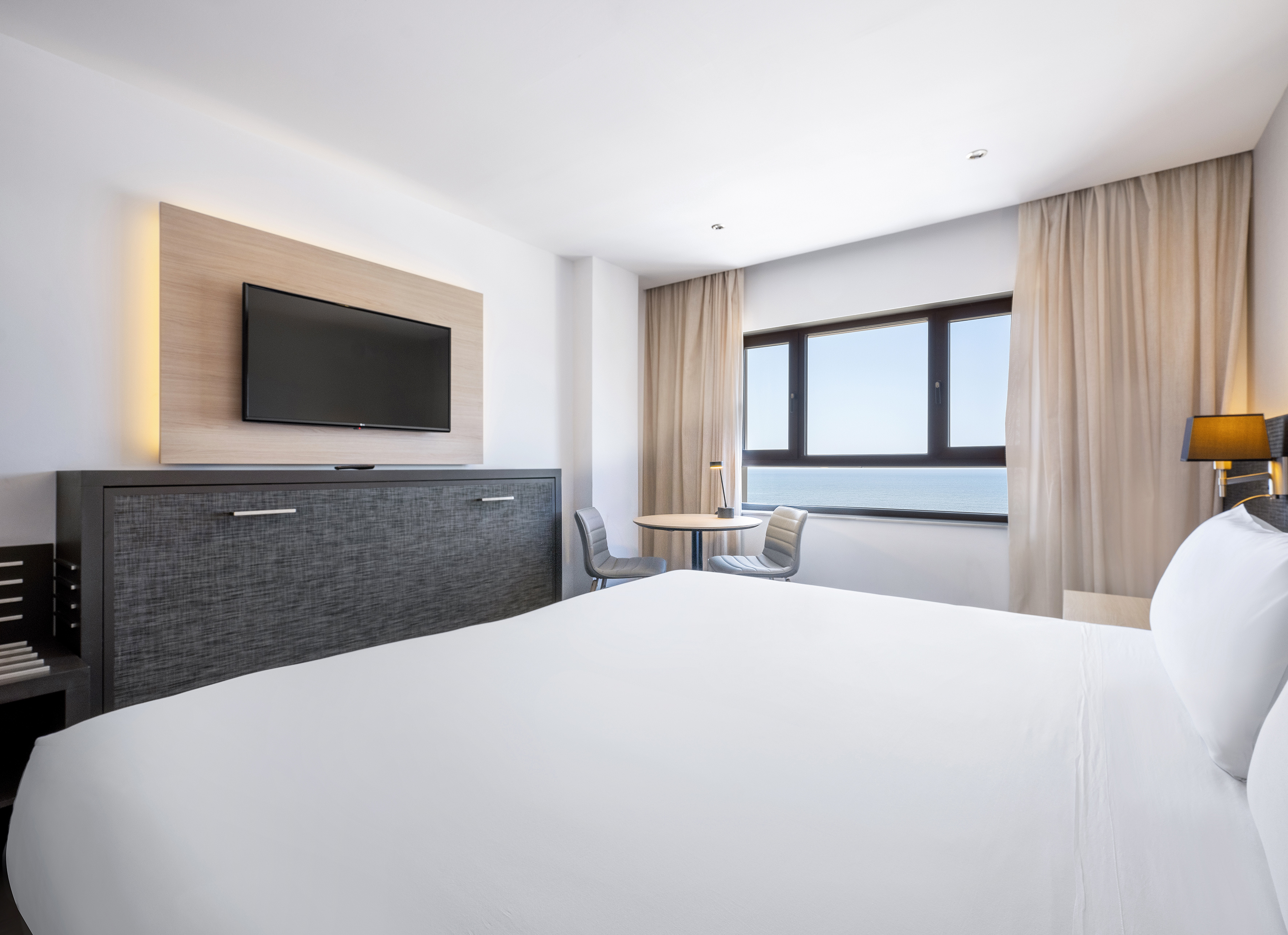 Modern ocean view hotel room with white bed, wall TV, and small table.