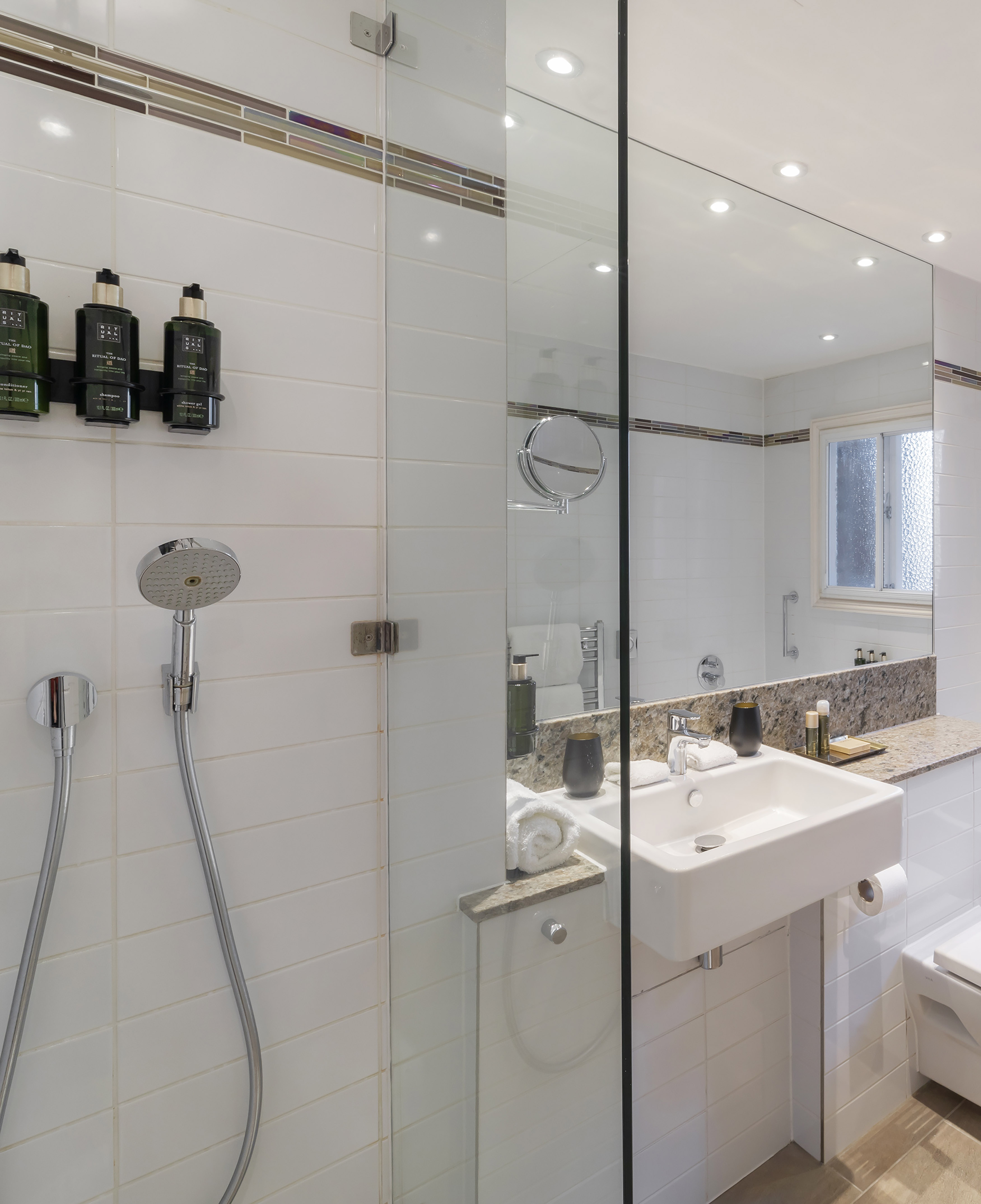 Hotel bathroom with white tiles, glass shower, Rituals products, and vanity.