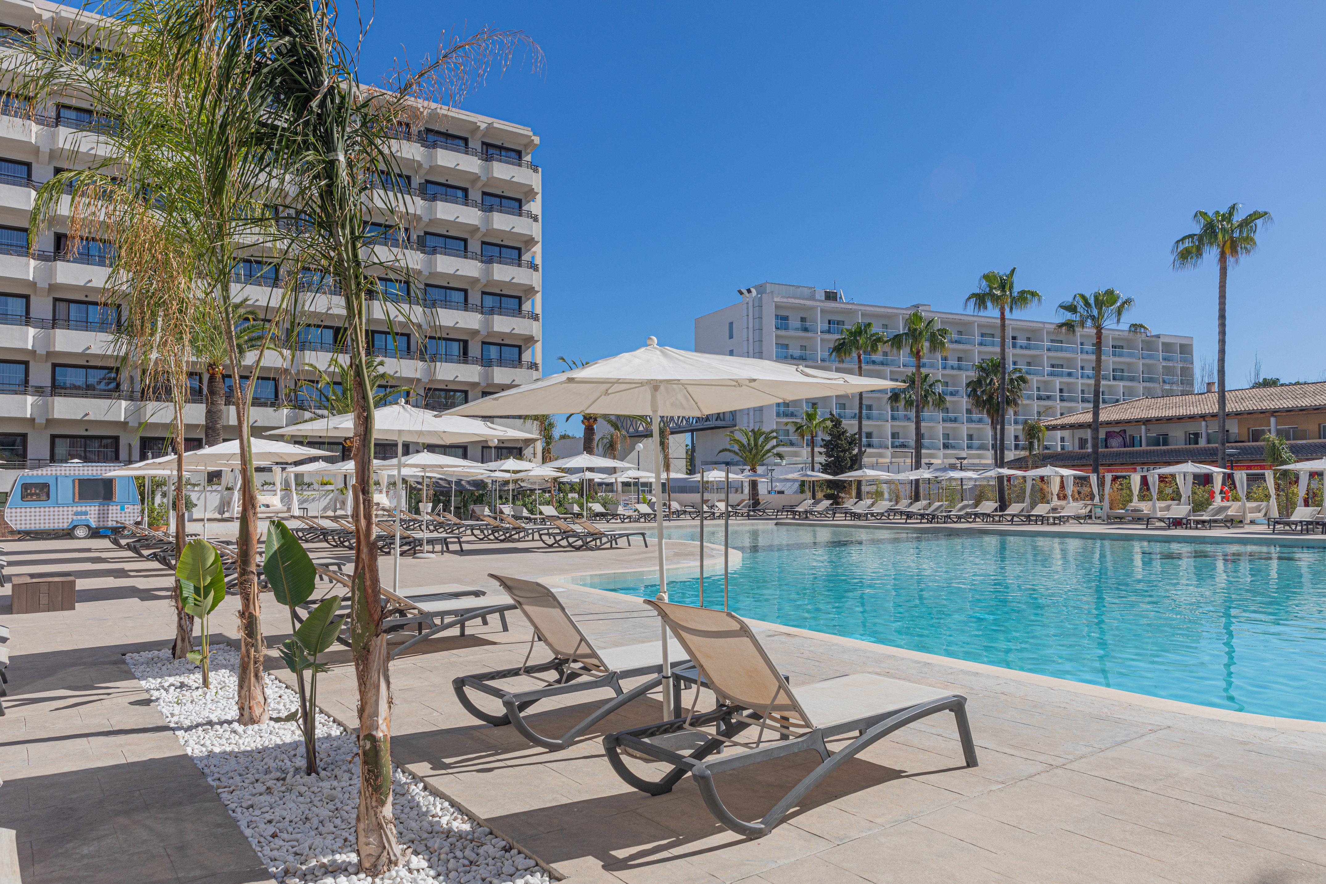 Sunny resort pool with sun loungers, palm trees, and modern hotel buildings.