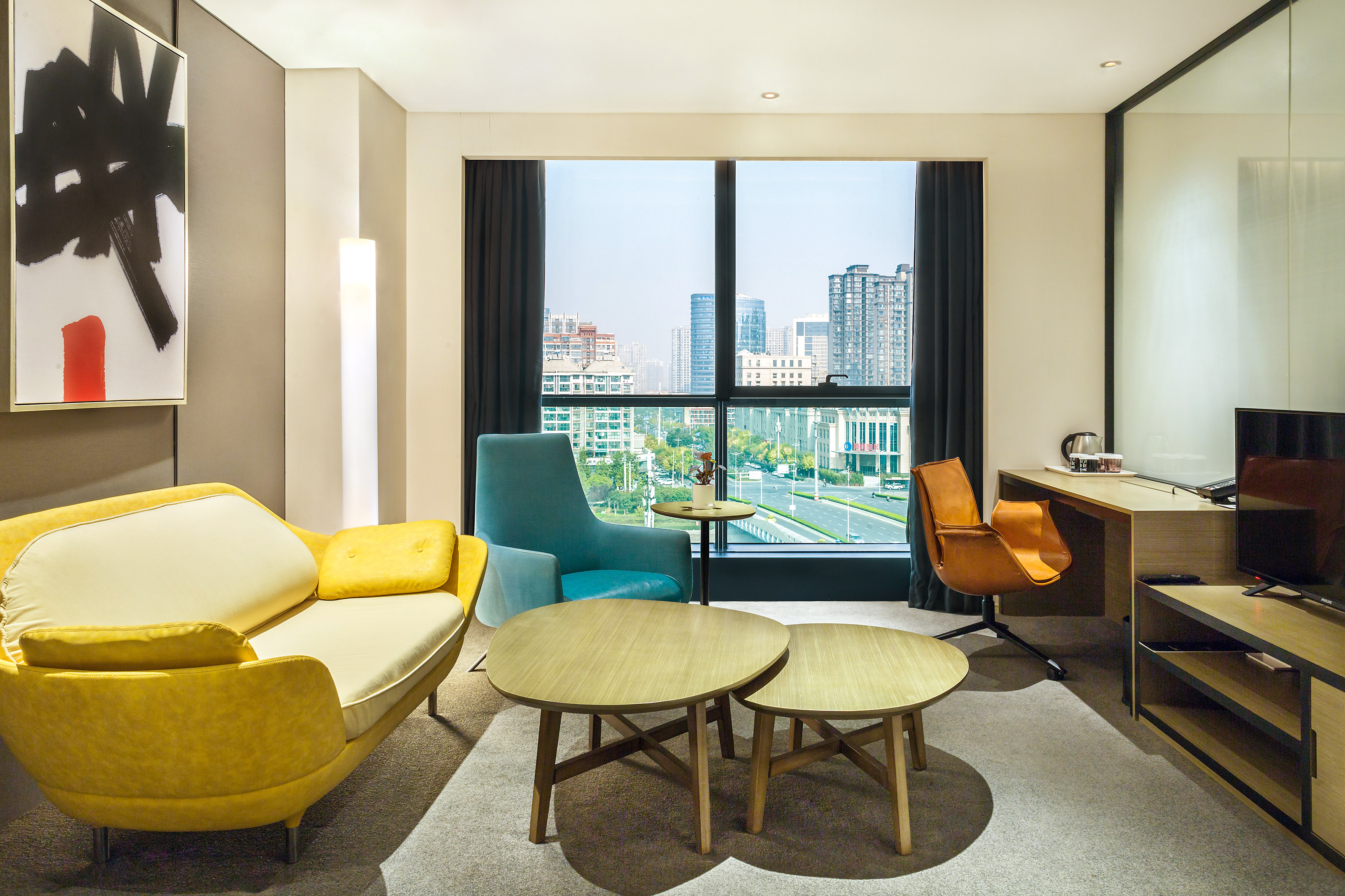 Modern hotel room living area with city view, yellow sofa, and work desk.
