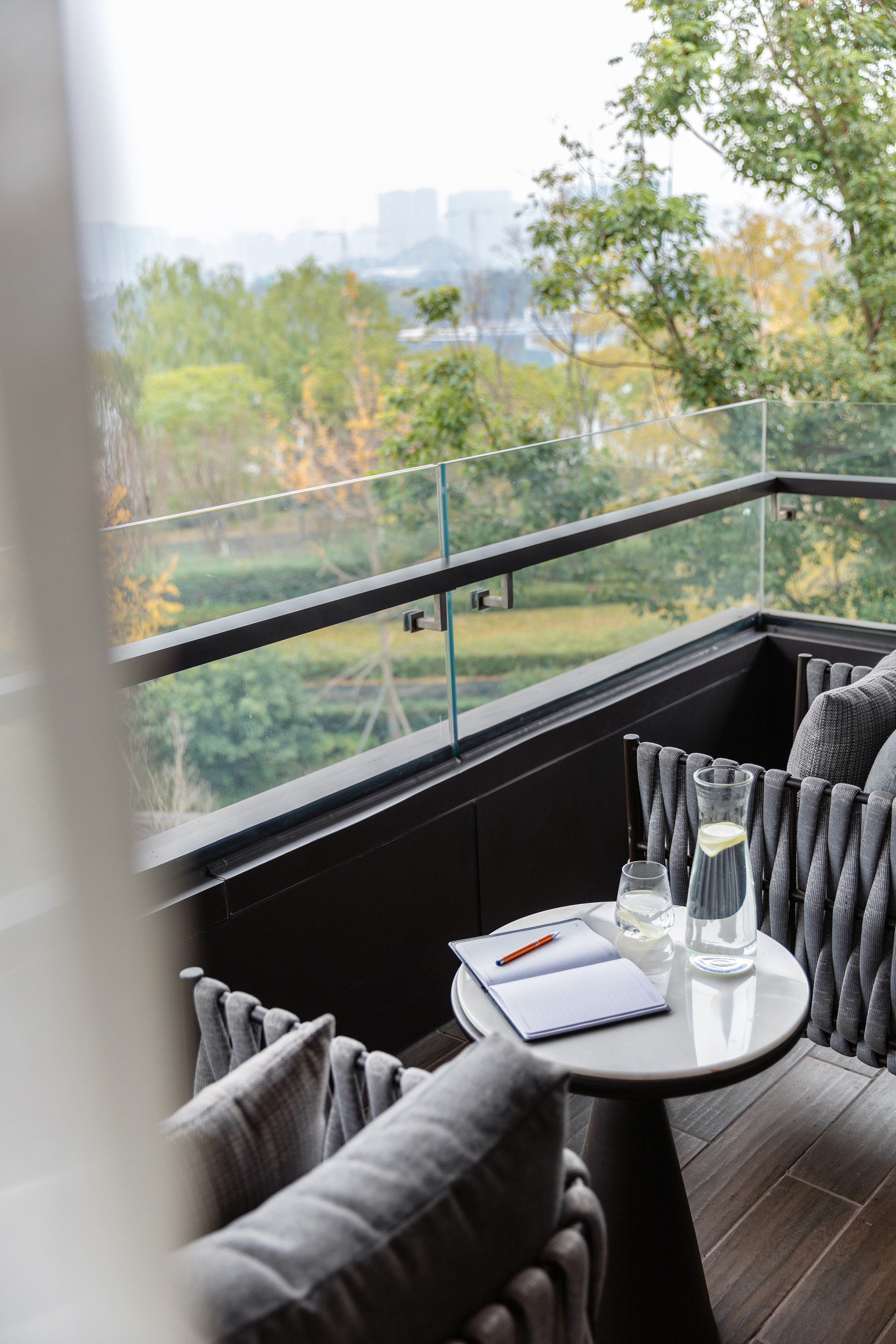 Modern balcony with outdoor furniture, notebook, water, and a city-forest view.