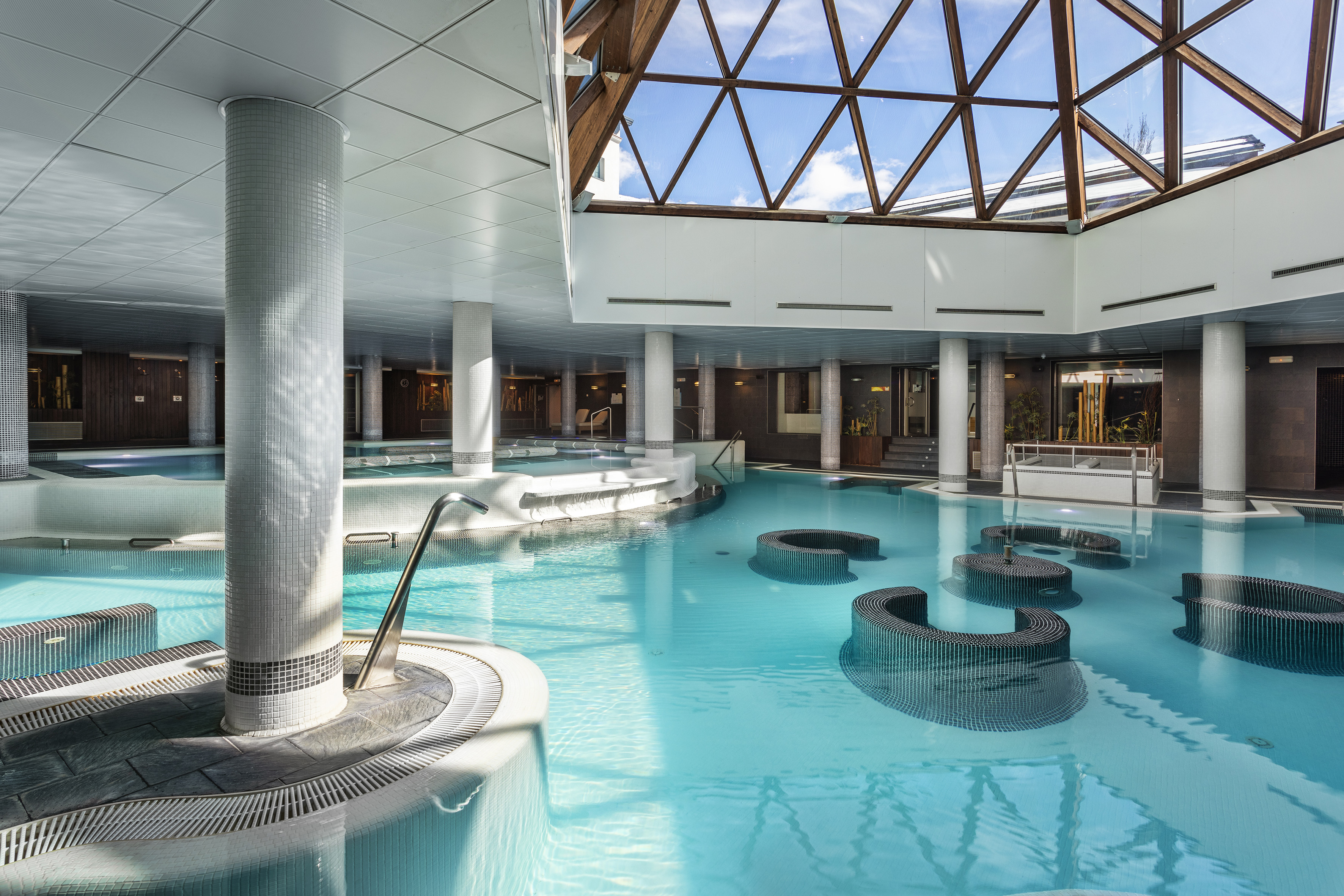 Bright indoor spa pool with glass roof, blue water, and hydrotherapy features.