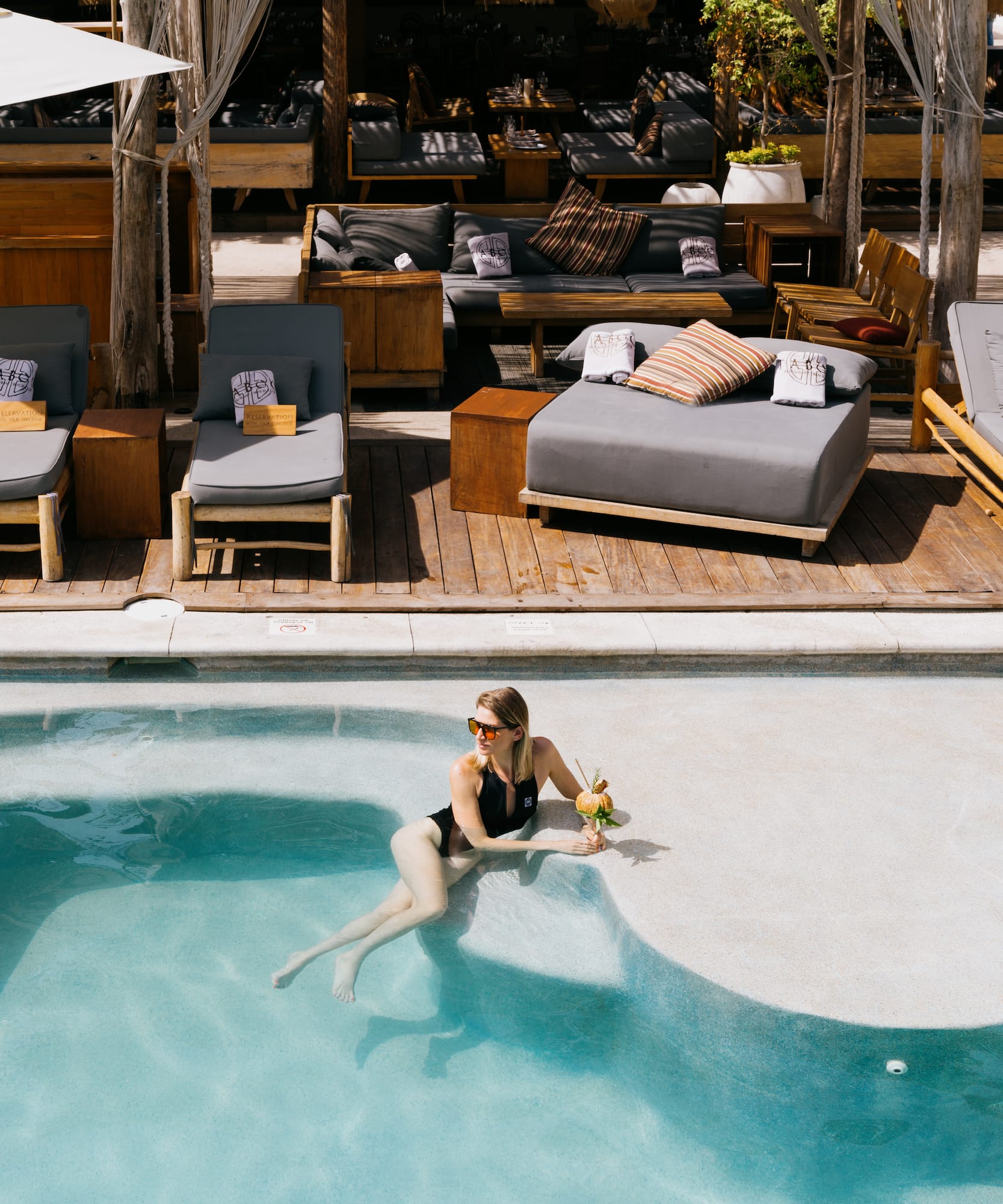 Woman in black swimsuit holding tropical drink in resort pool, lounge area.