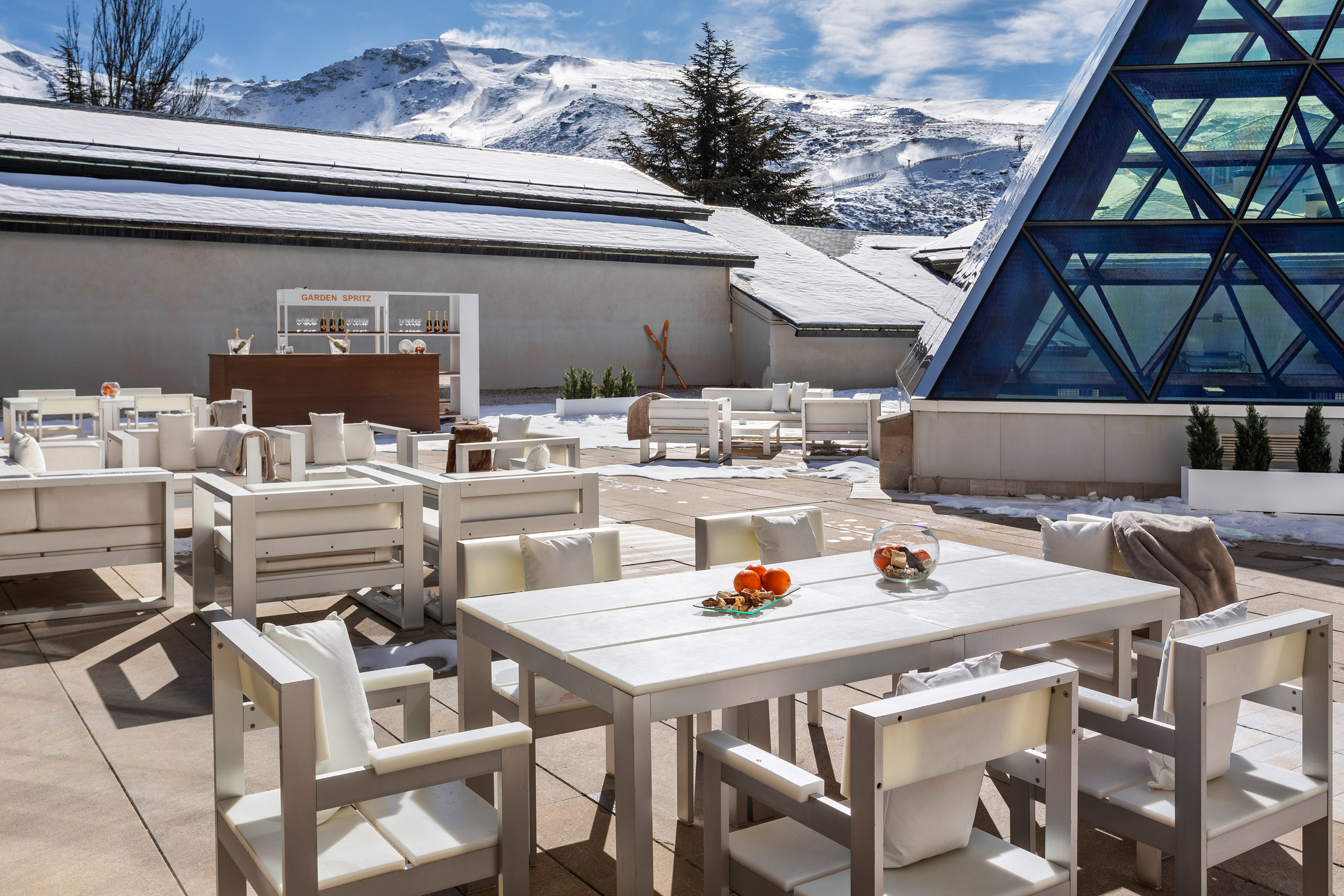 Ski resort terrace with white lounge furniture, outdoor bar, snowy mountains.