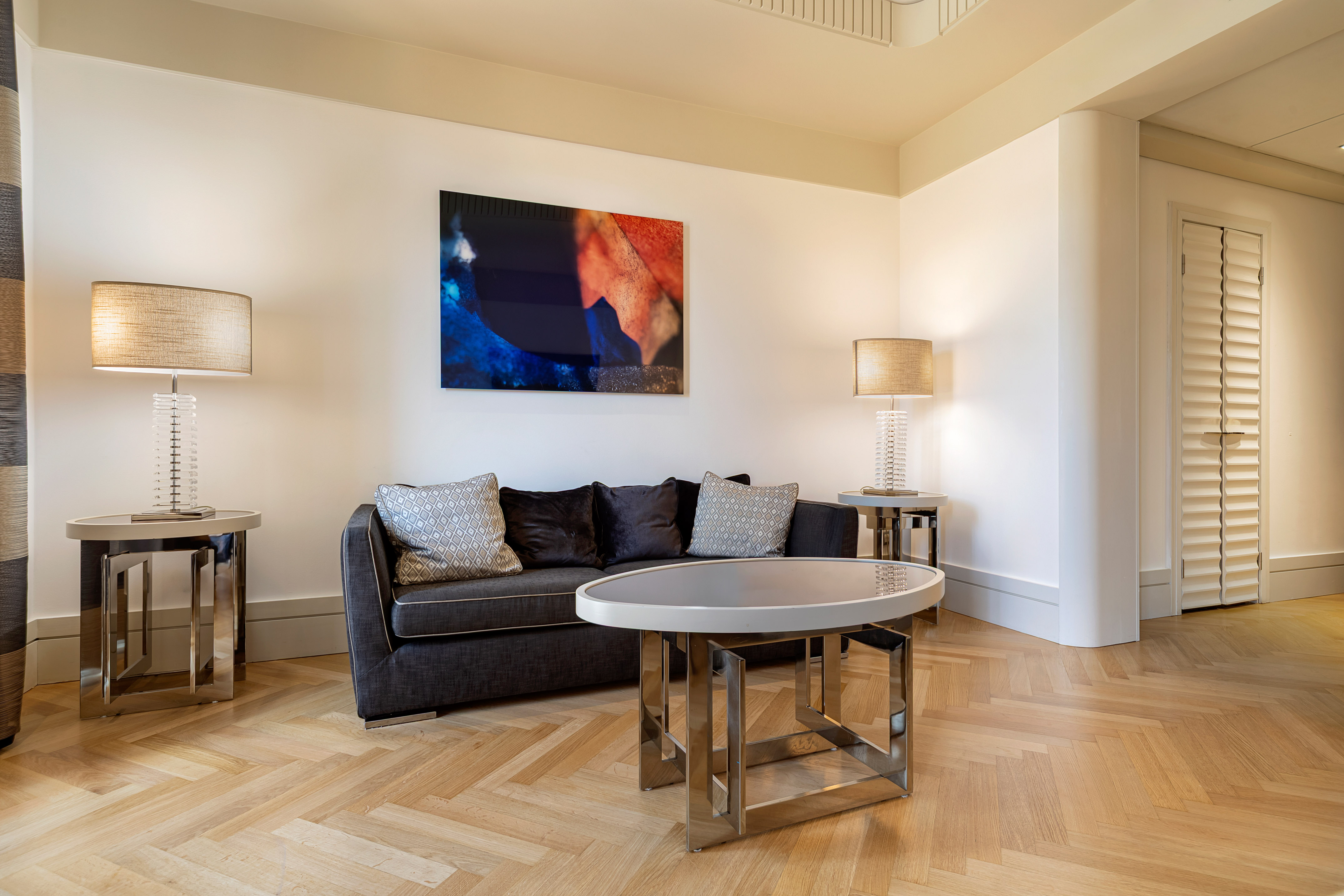 Modern living room with dark sofa, abstract art, two lamps, and herringbone wood floor.