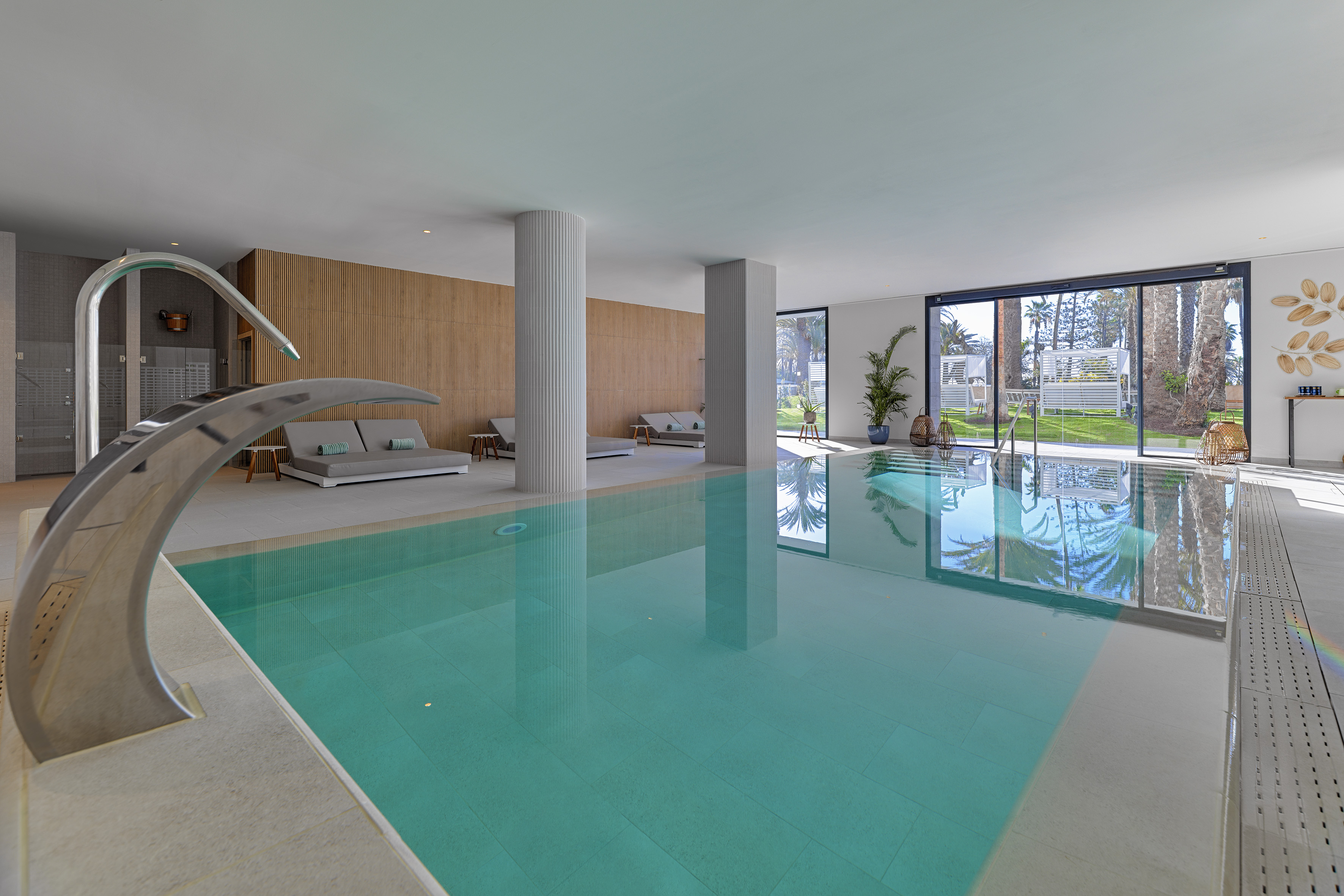 Modern indoor swimming pool with turquoise water, lounge daybeds, and palm tree view.