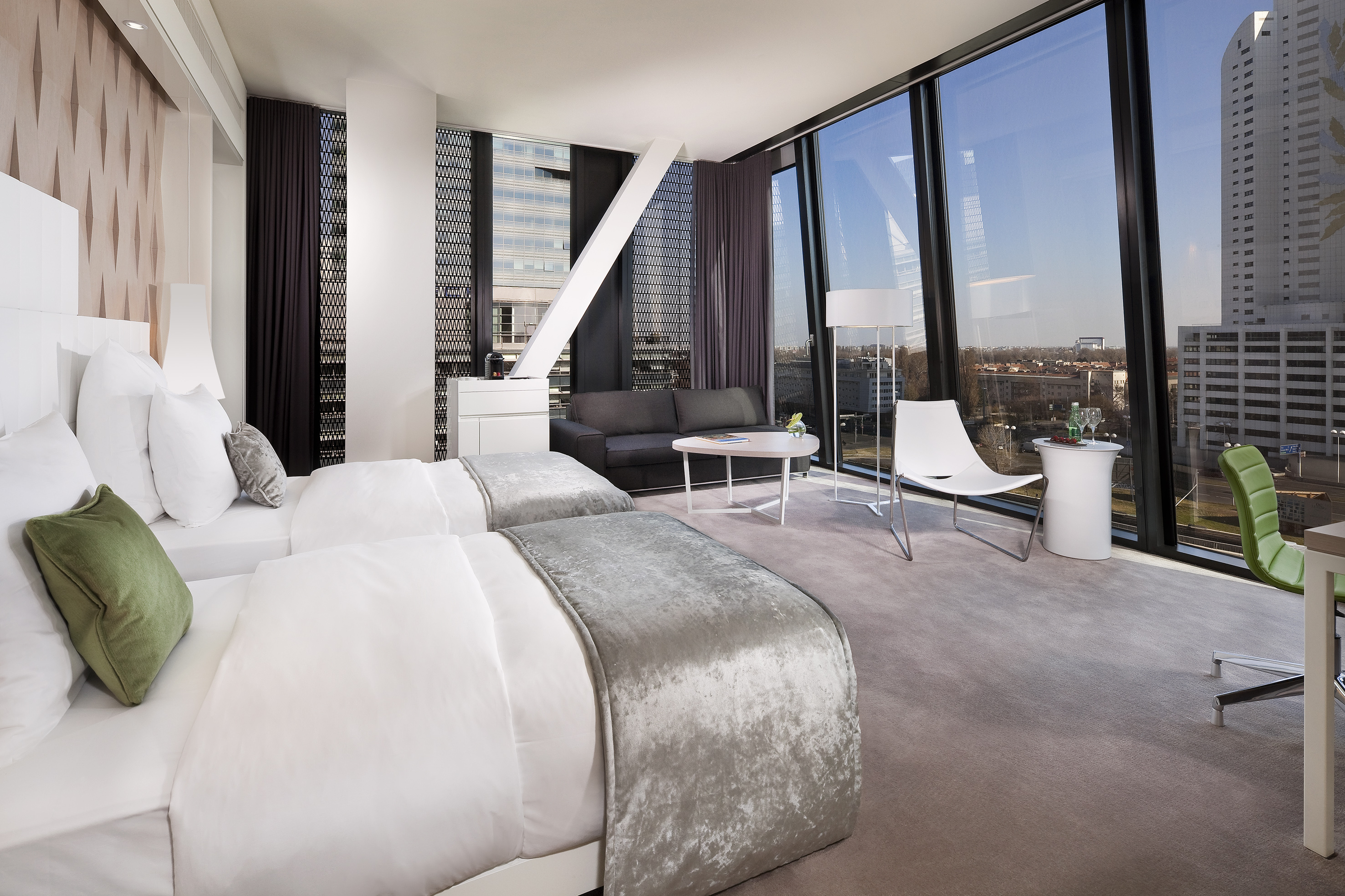 Modern hotel room with twin beds, sofa, and floor-to-ceiling city view windows.