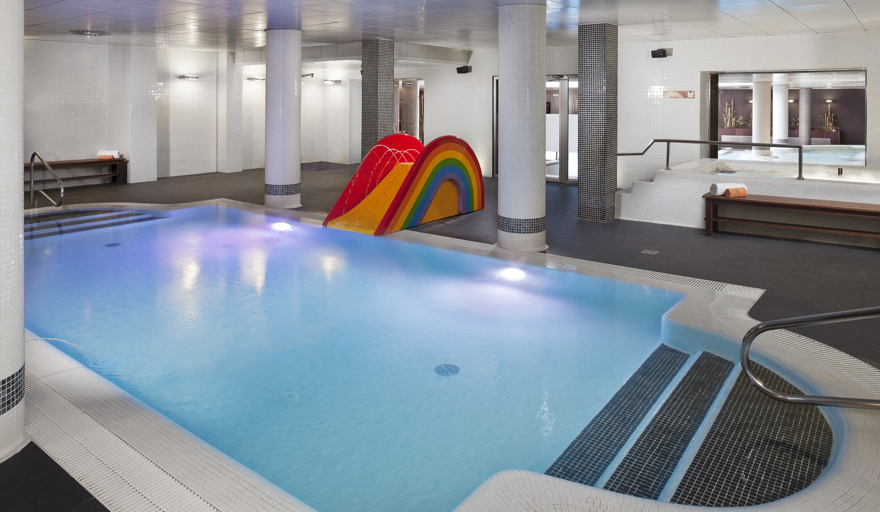 Indoor swimming pool with stairs, a colorful rainbow slide, and spa area.