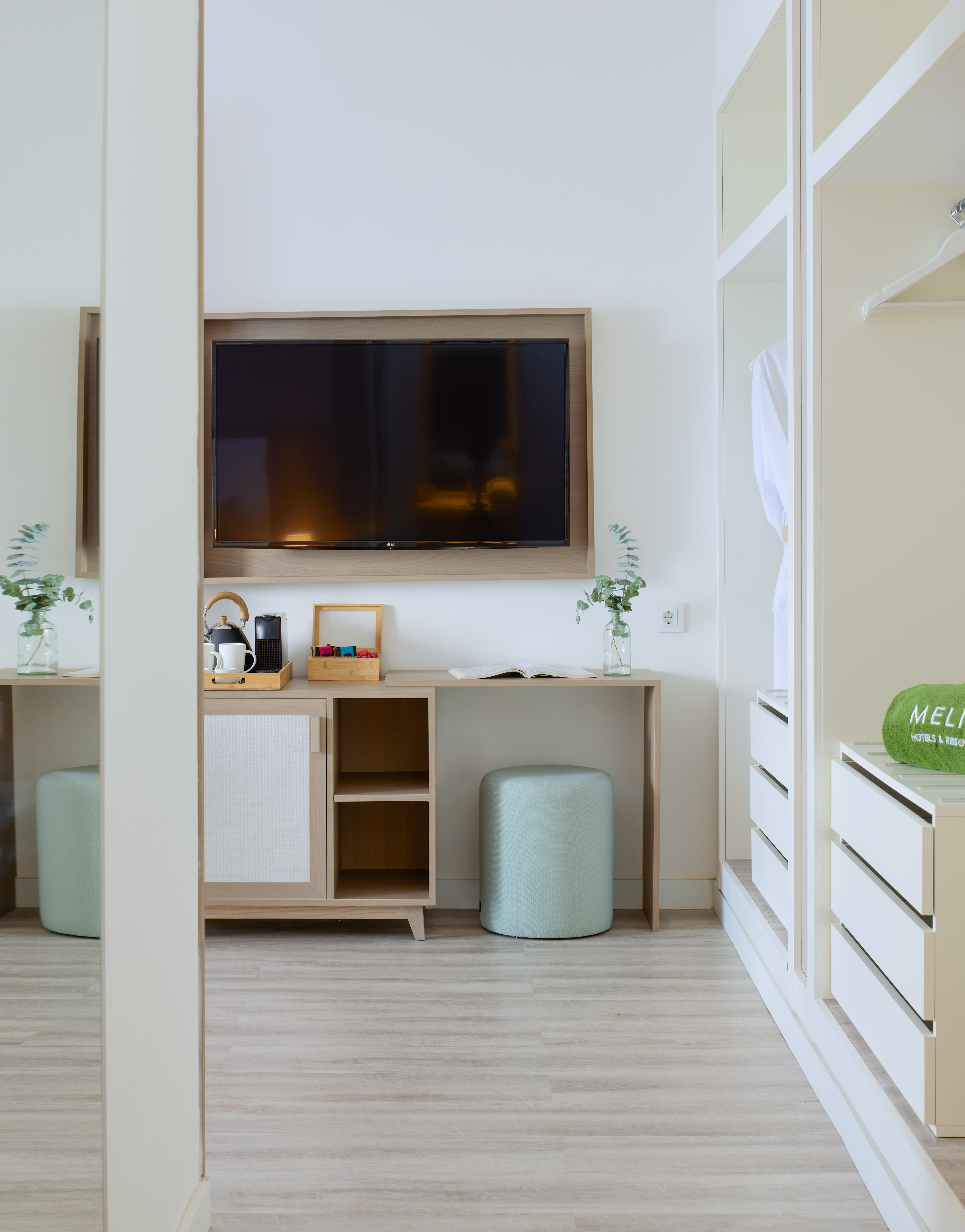 Modern hotel room with TV, coffee station, blue ottoman, and open wardrobe.