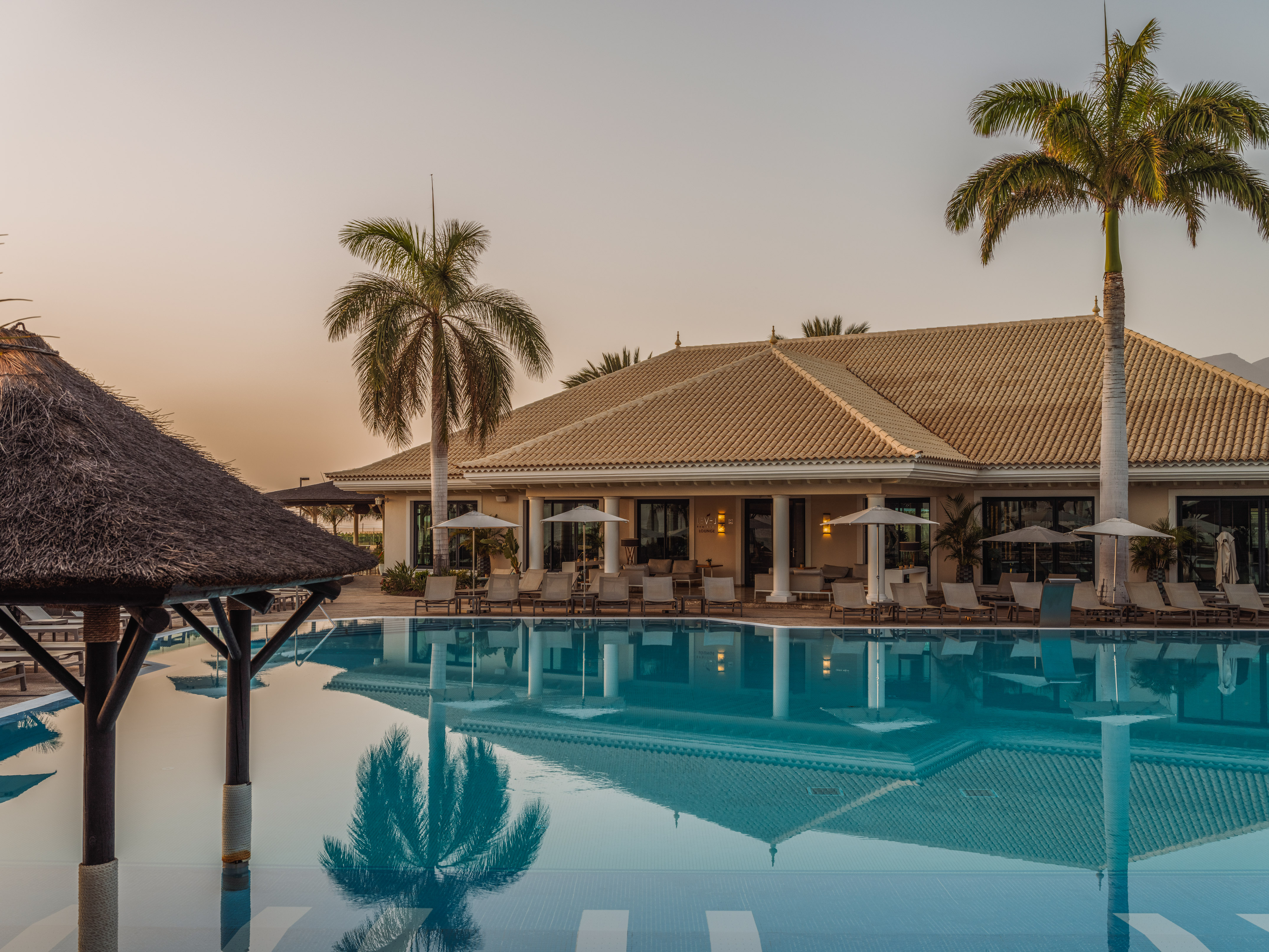 Tropical resort pool with palm trees, lounge chairs, and reflections at sunset.