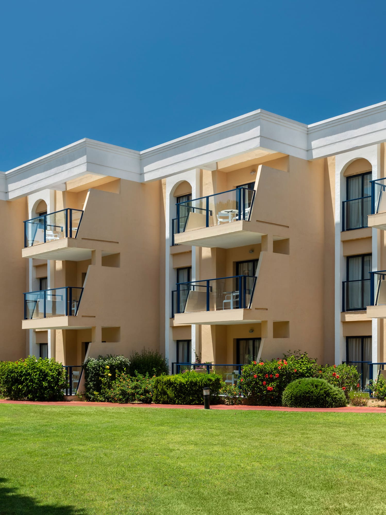 Sunny hotel building with balconies and a lush green lawn.