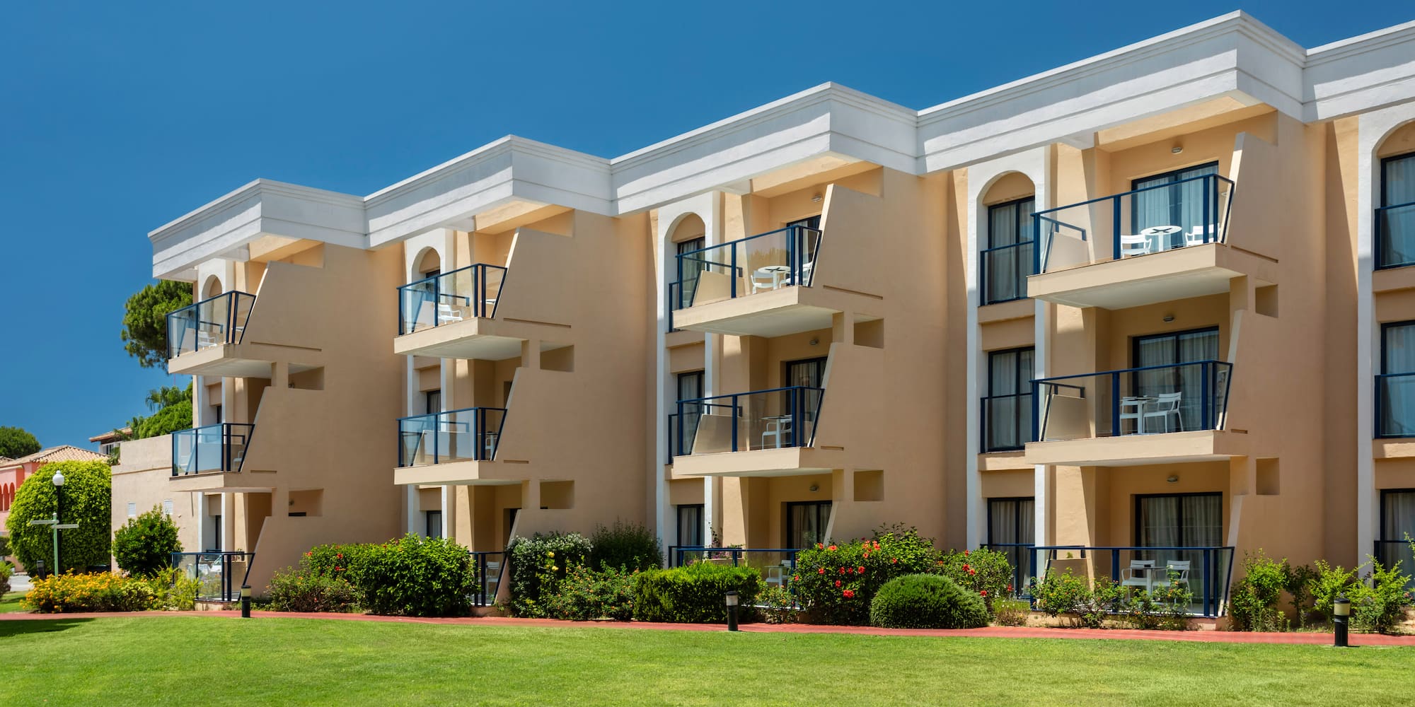 Sunny resort building with multiple balconies, green lawn, and garden landscaping.