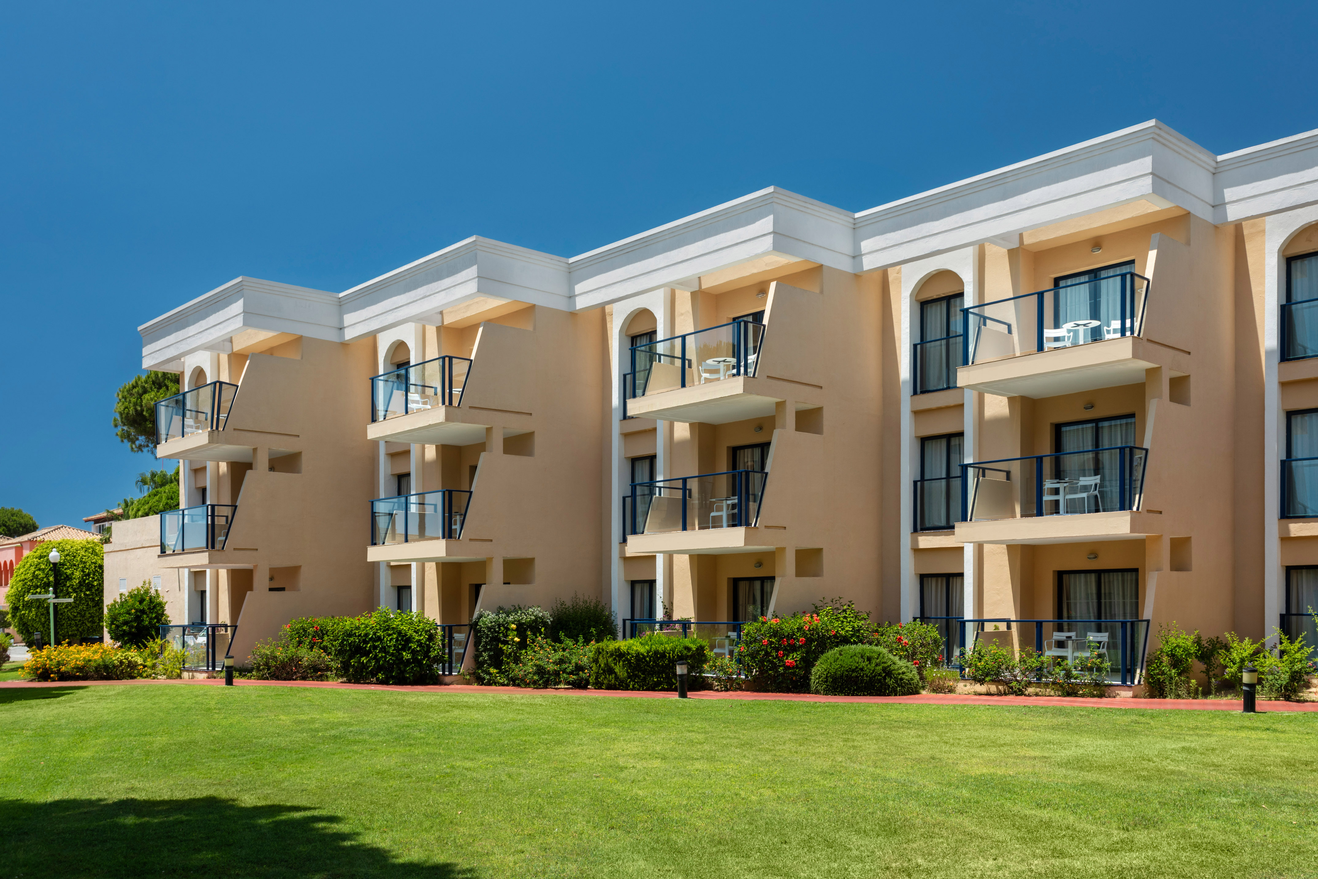 Sunny hotel building with balconies and a lush green lawn.