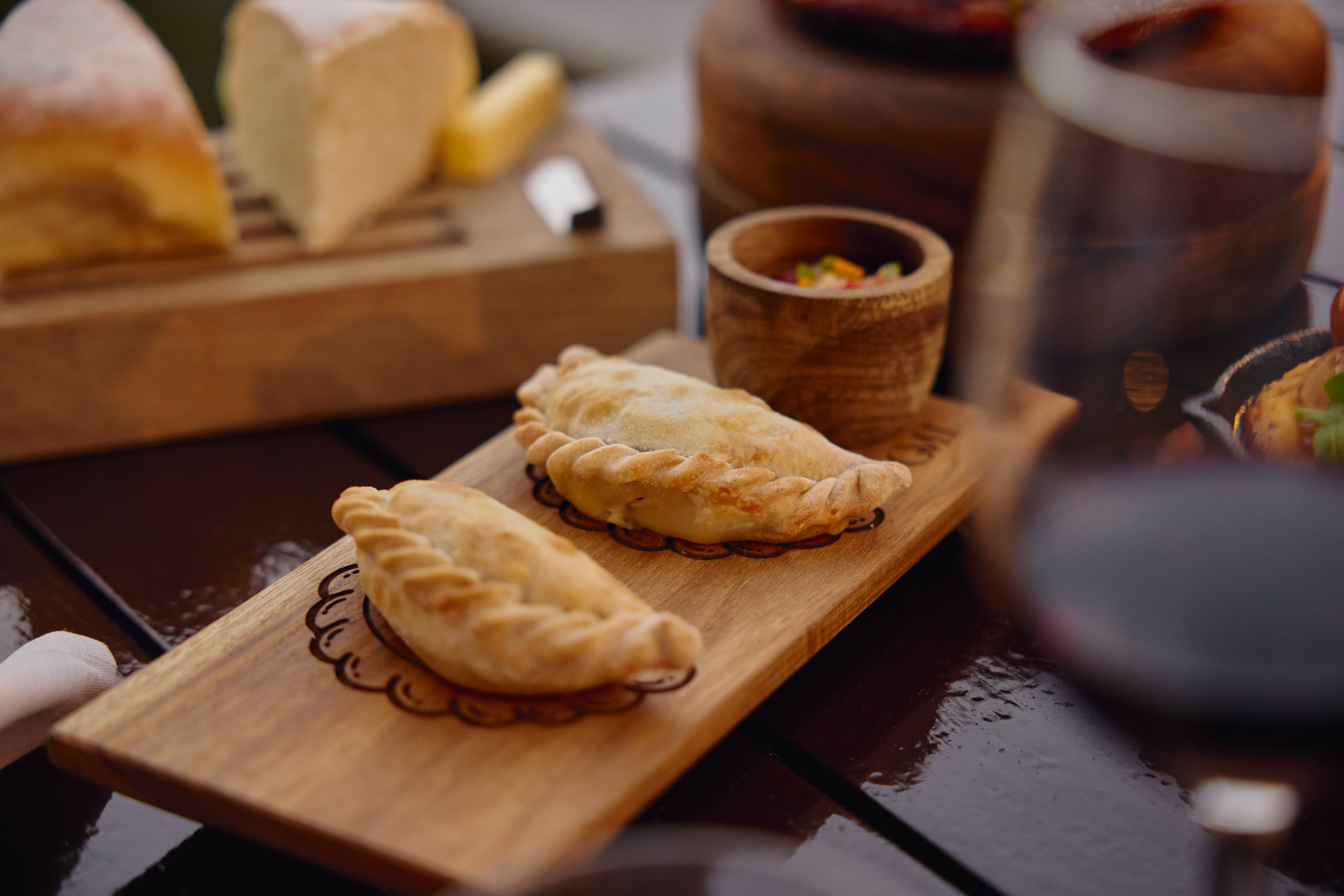 Empanadas doradas sobre tabla de madera, con salsa, queso y vino.