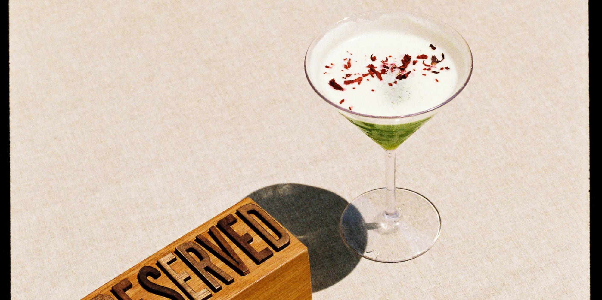 Wooden RESERVED sign and green cocktail with red garnish on a light table.