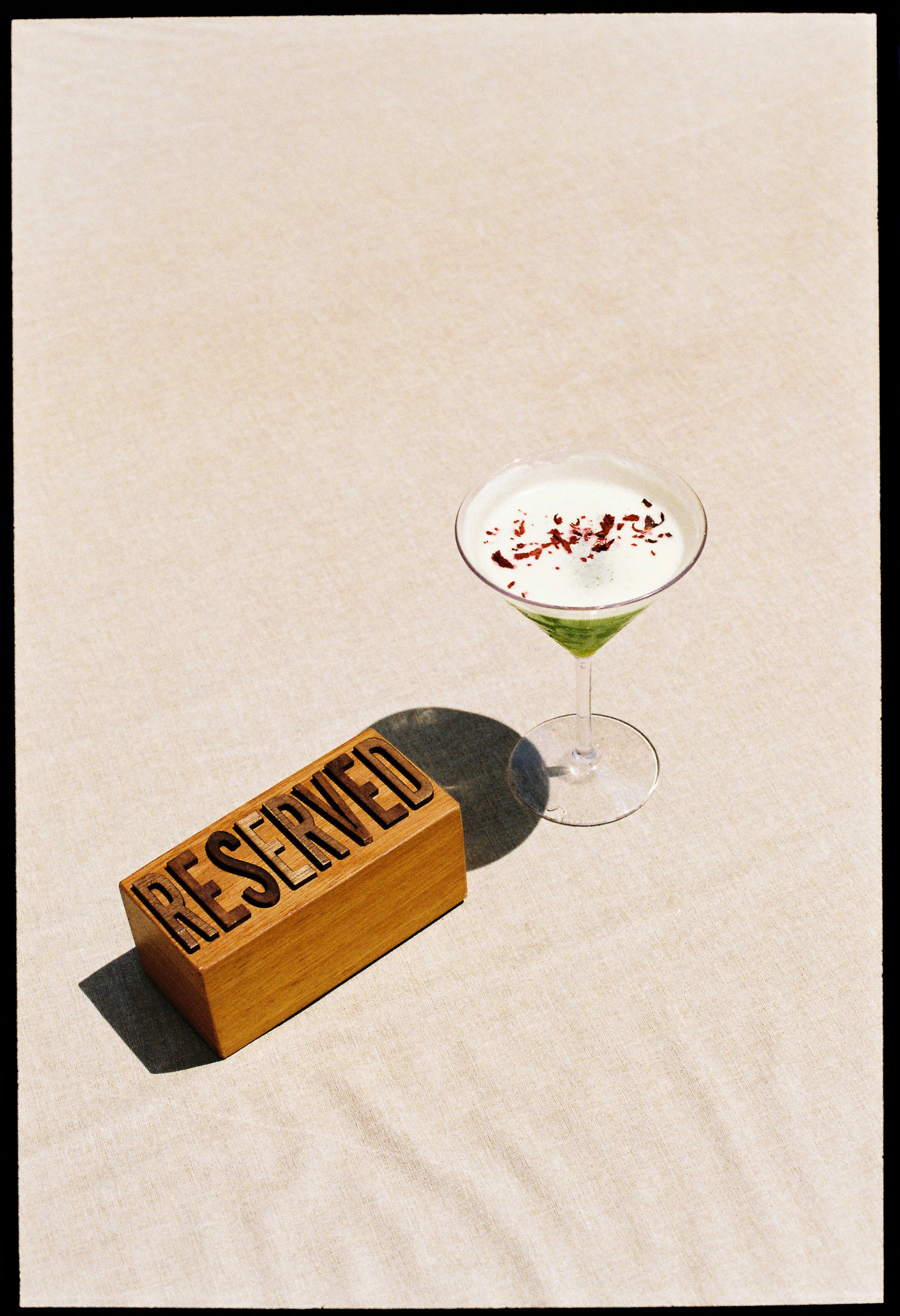 Wooden RESERVED sign and green cocktail with red garnish on a light table.