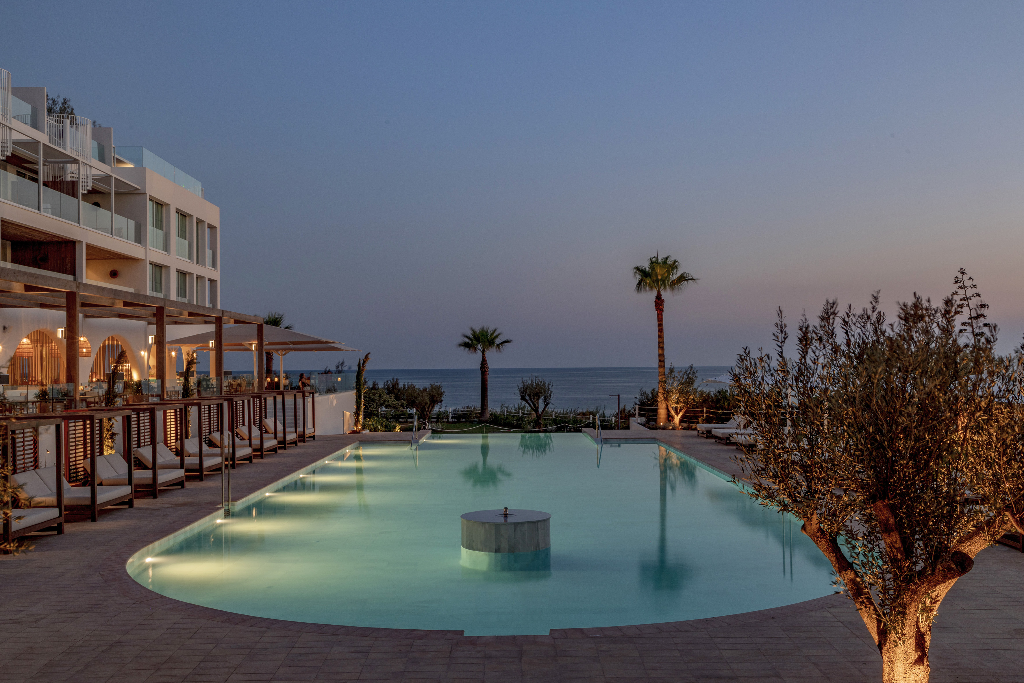 Illuminated resort swimming pool with ocean view and palm trees at dusk.
