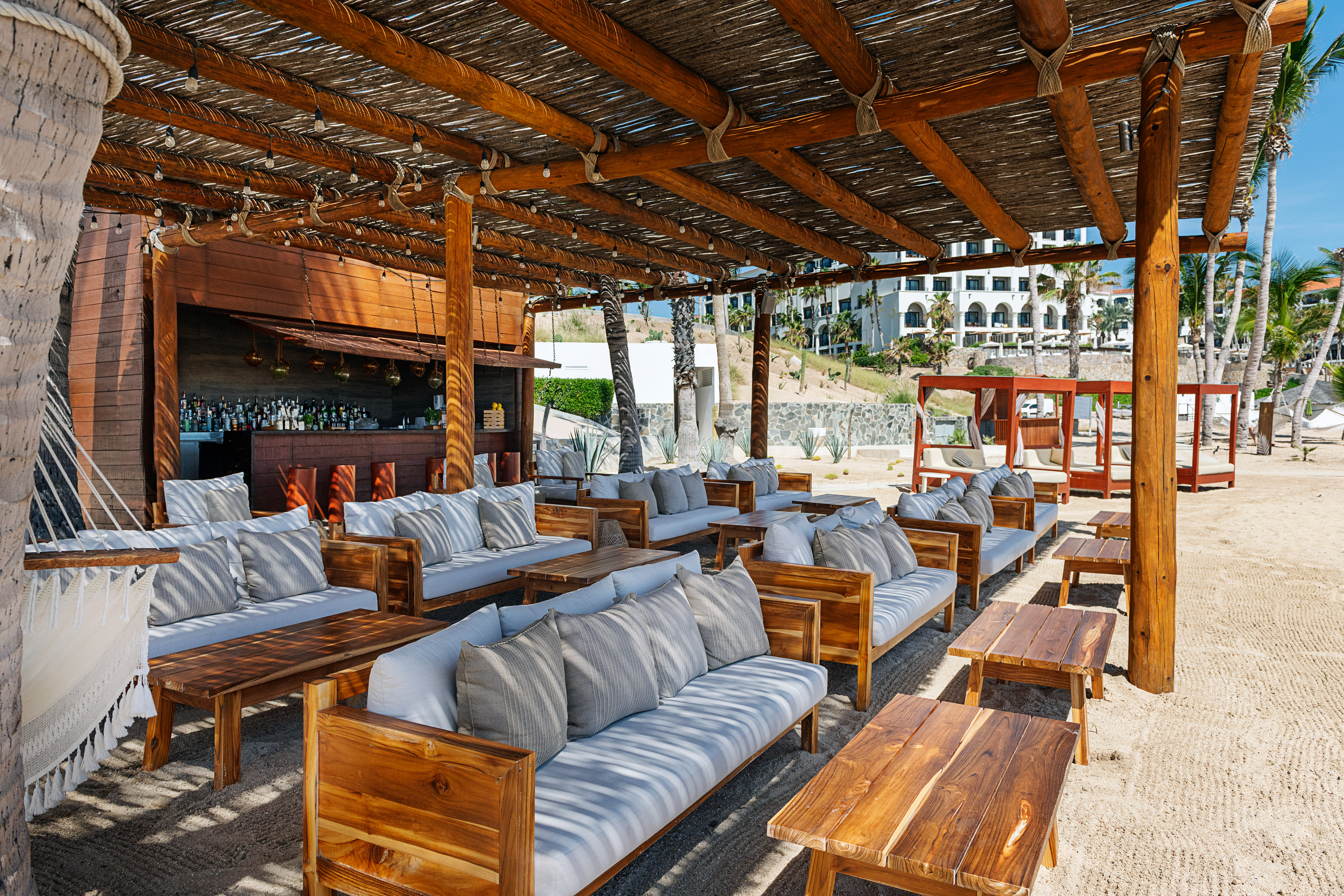 Beach bar and resort lounge with wooden sofas, thatched roof, on sand.