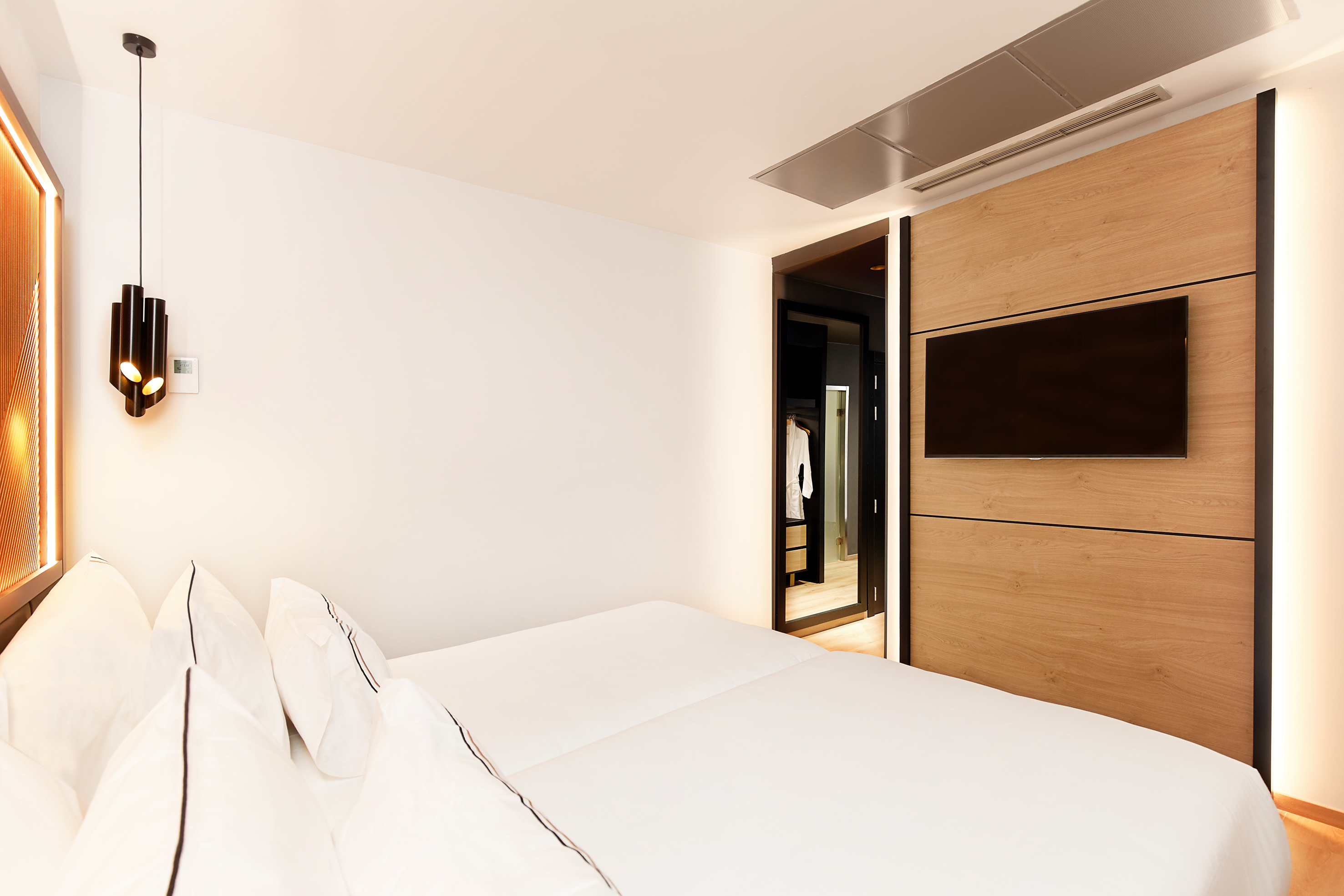 Modern hotel bedroom with white bed, wood TV panel, and sleek lighting.
