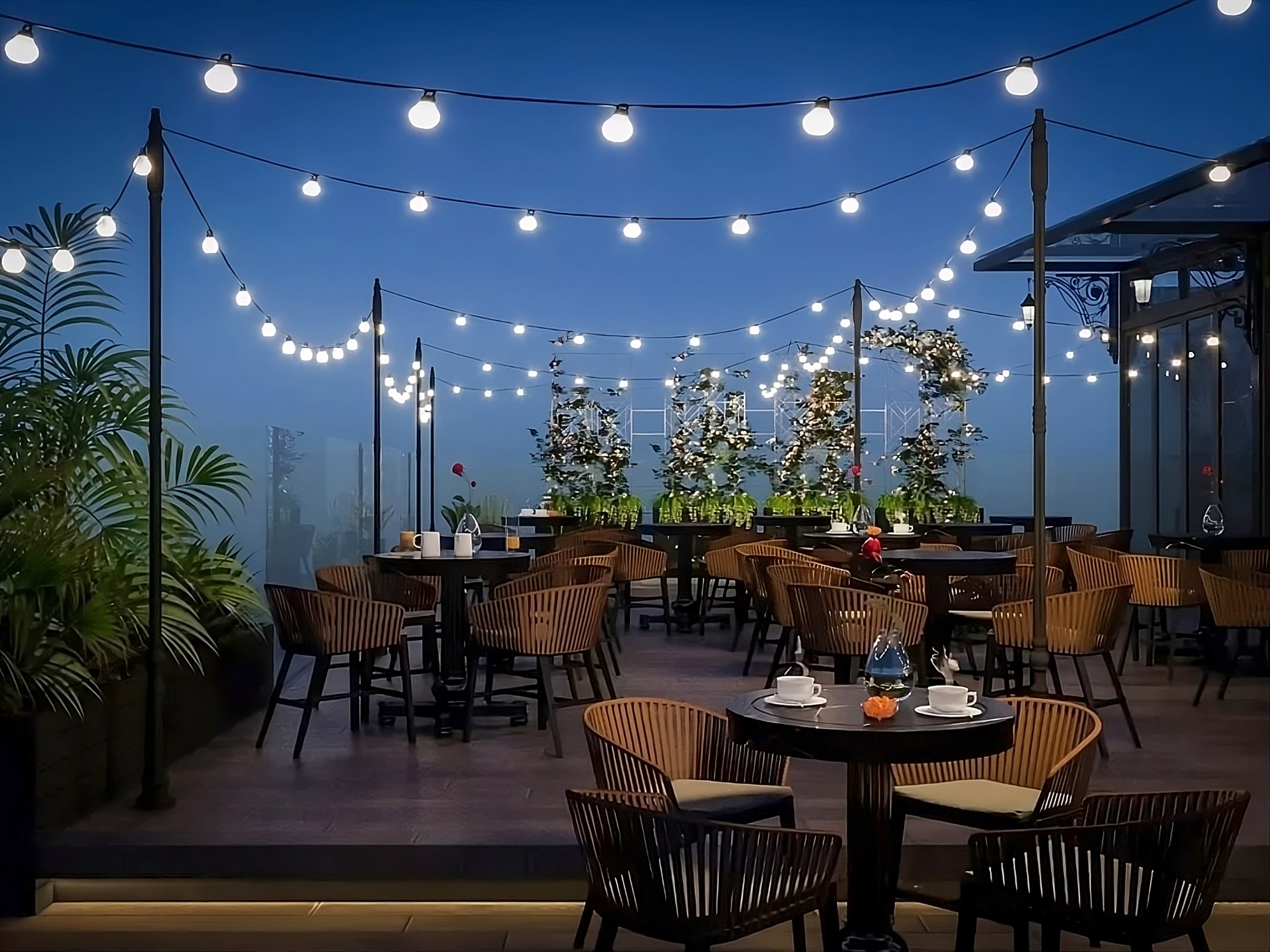 Rooftop dining patio with string lights, dark tables, and wooden chairs at dusk.
