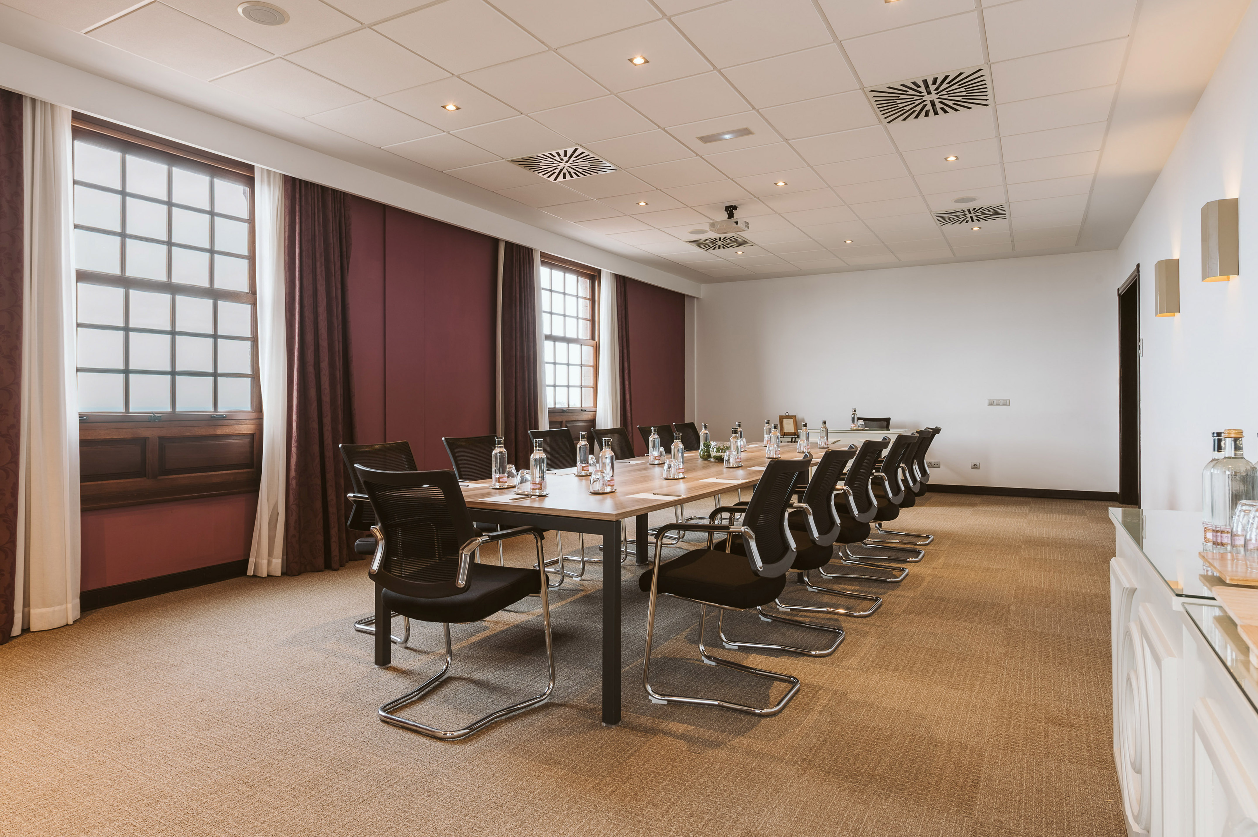 Conference room with long table, black chairs, water bottles, and tall windows.
