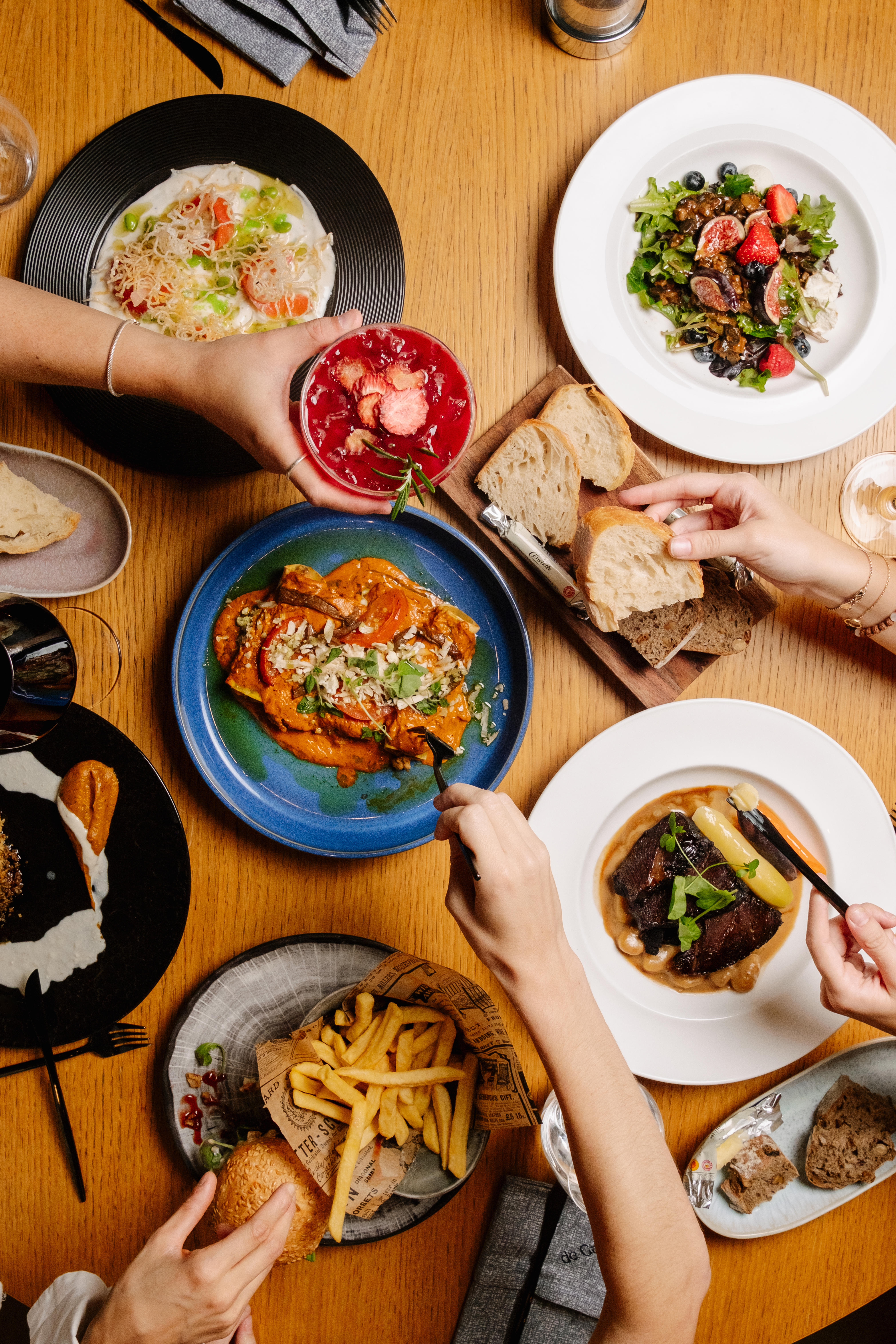 Overhead view: diverse restaurant dishes with hands dining on a wooden table.