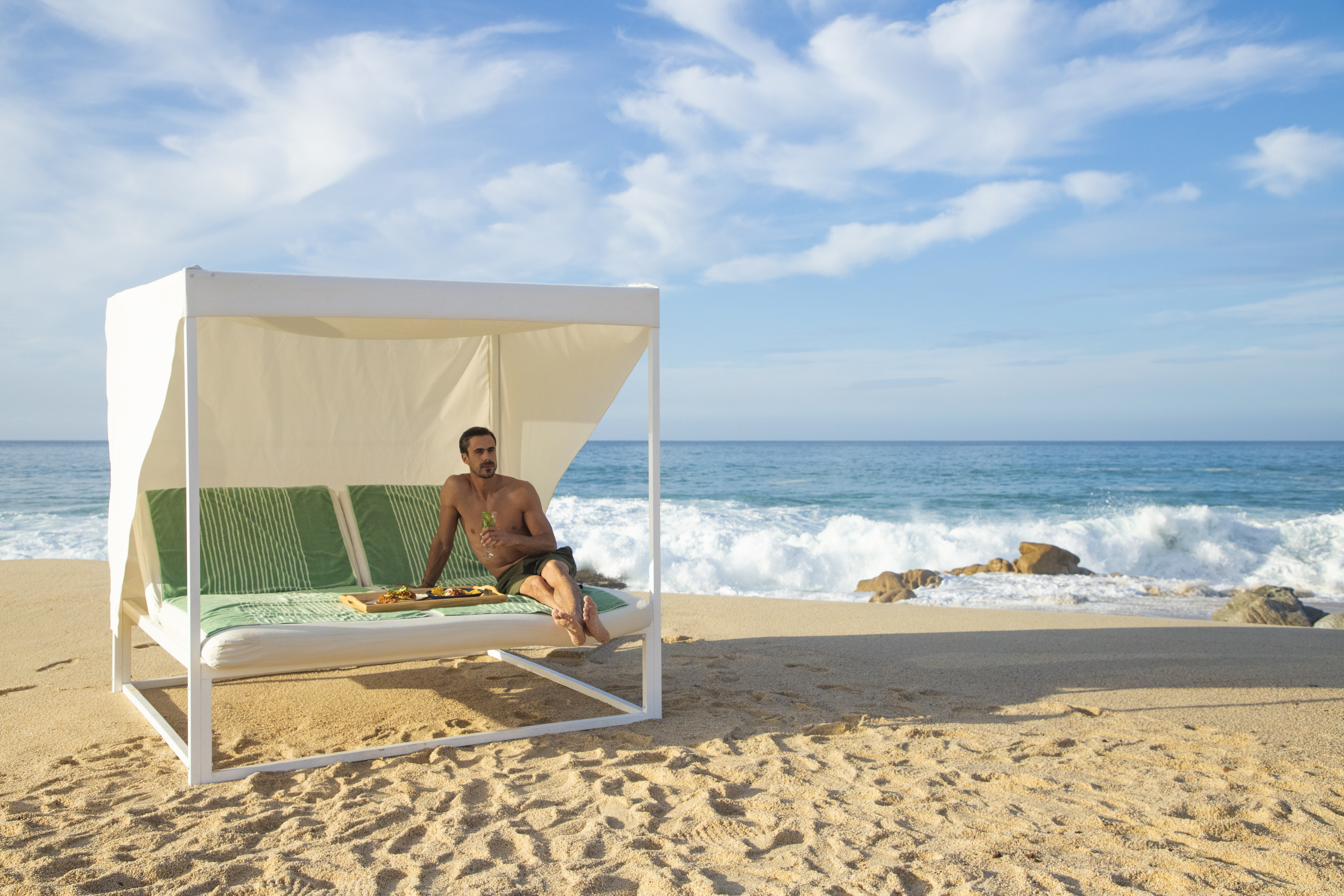 Man relaxing on beach cabana with drink, food tray, ocean waves.