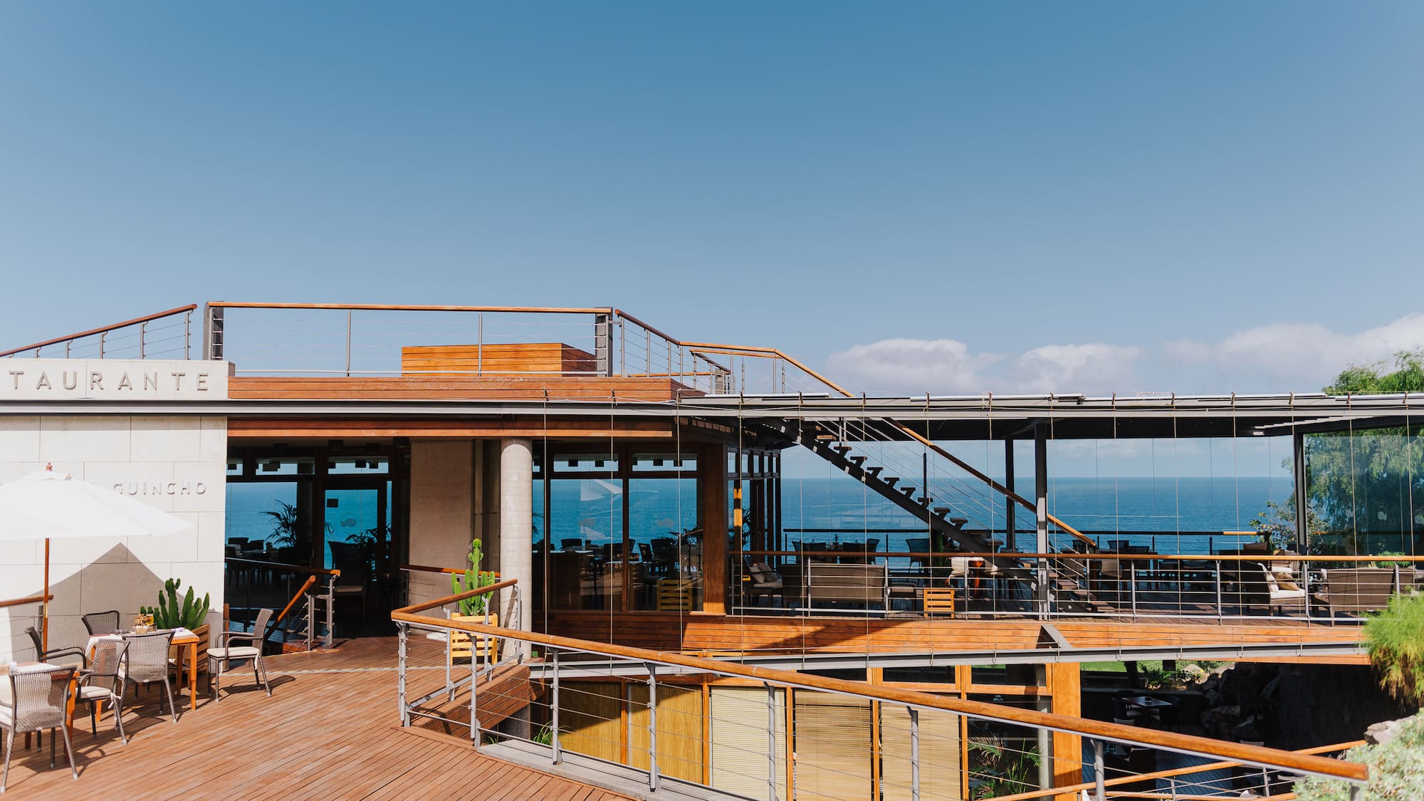 Multi-level oceanfront restaurant with wooden decks, glass walls, outdoor dining, and blue sky.