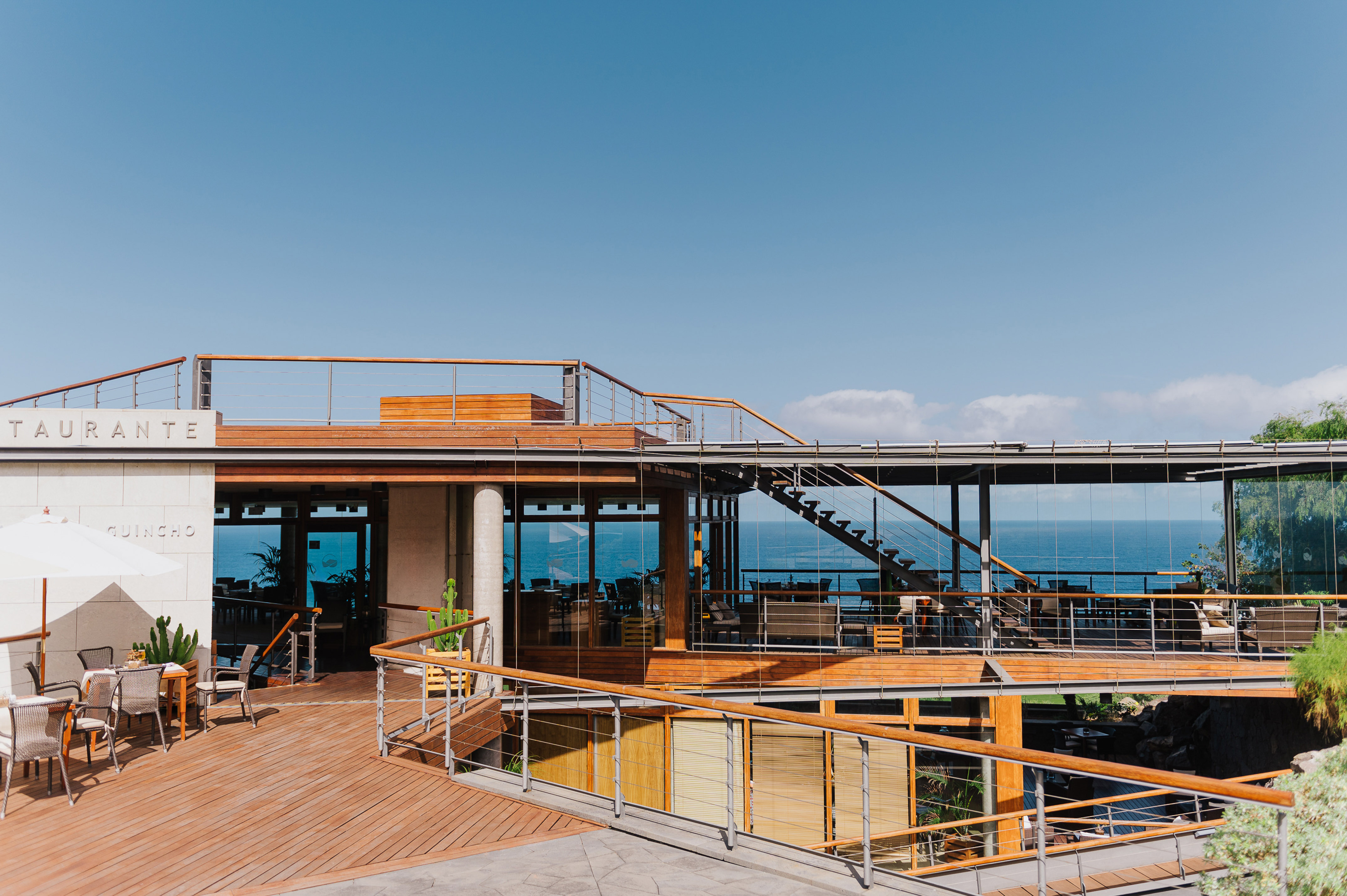 Multi-level oceanfront restaurant with wooden decks, glass walls, outdoor dining, and blue sky.