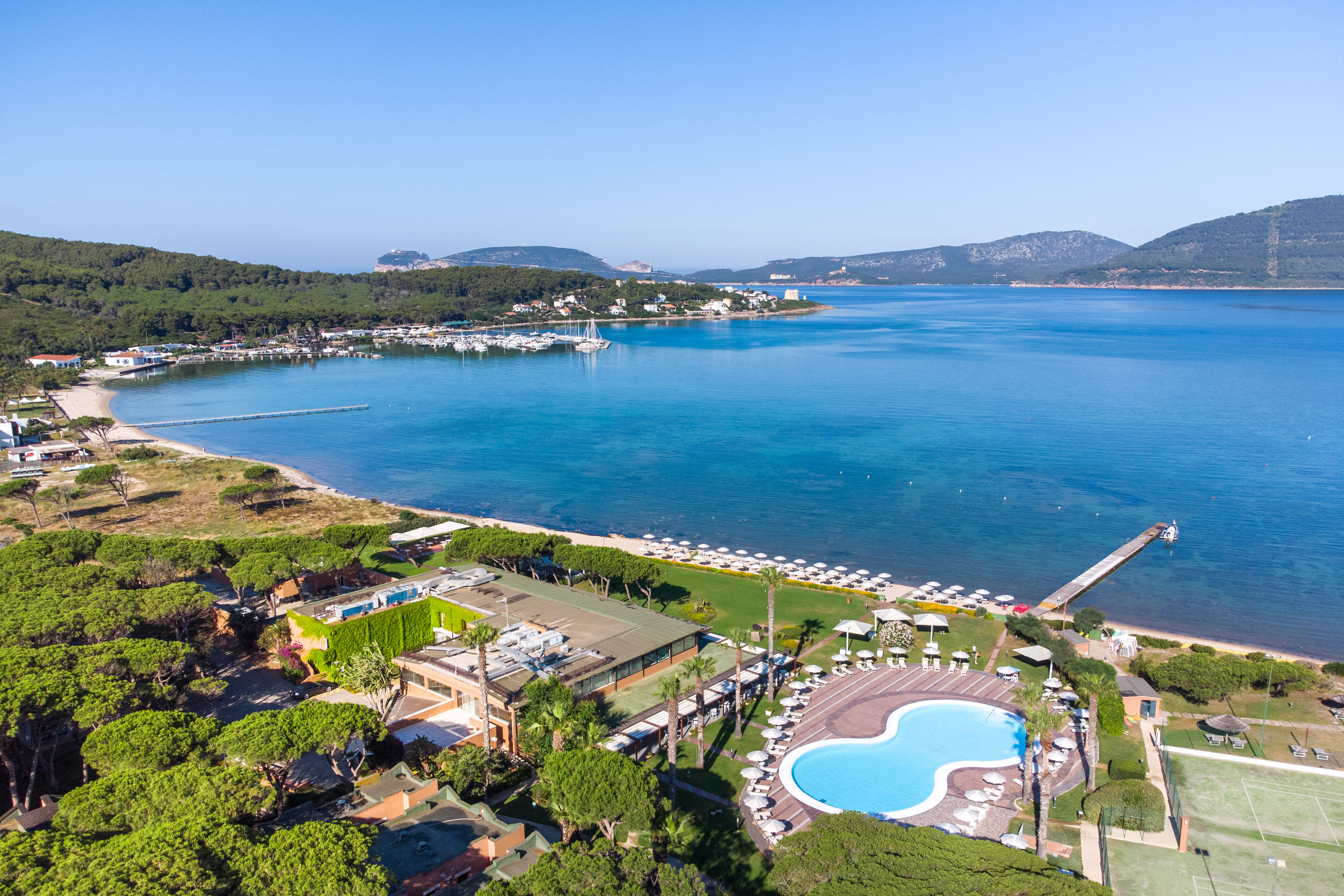 Aerial view: coastal resort, large swimming pool, sunny beach, marina, and green bay.
