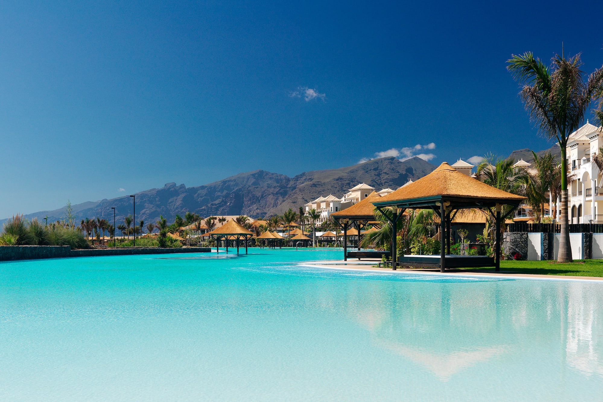 Resort tropical con laguna azul, cabañas, palmeras y montañas.
