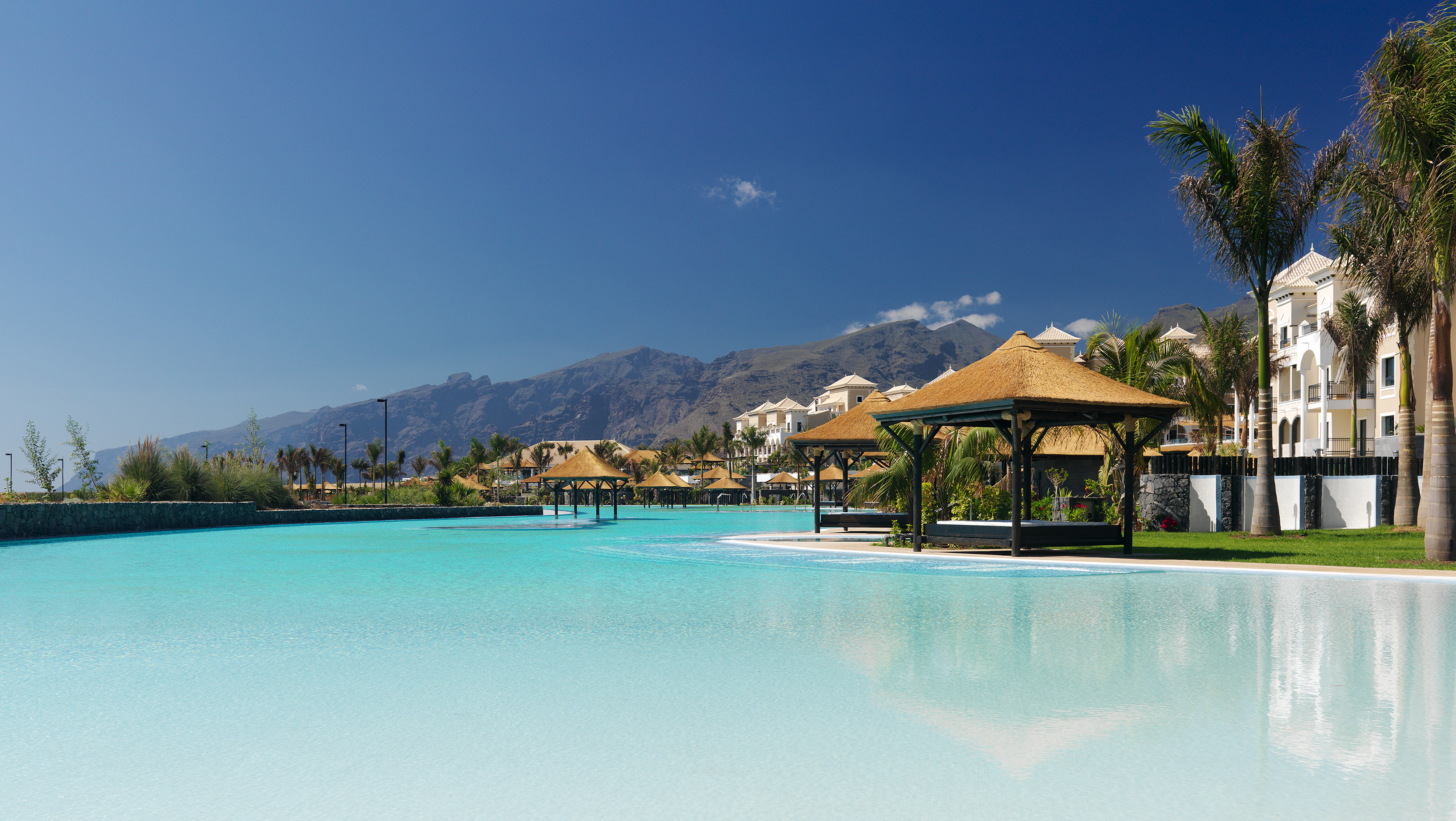 Resort tropical con laguna azul, cabañas, palmeras y montañas.