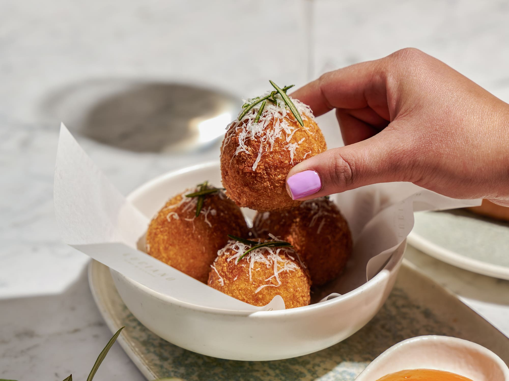 Hand lifting golden fried arancini with parmesan and rosemary from bowl, wine, sauce.