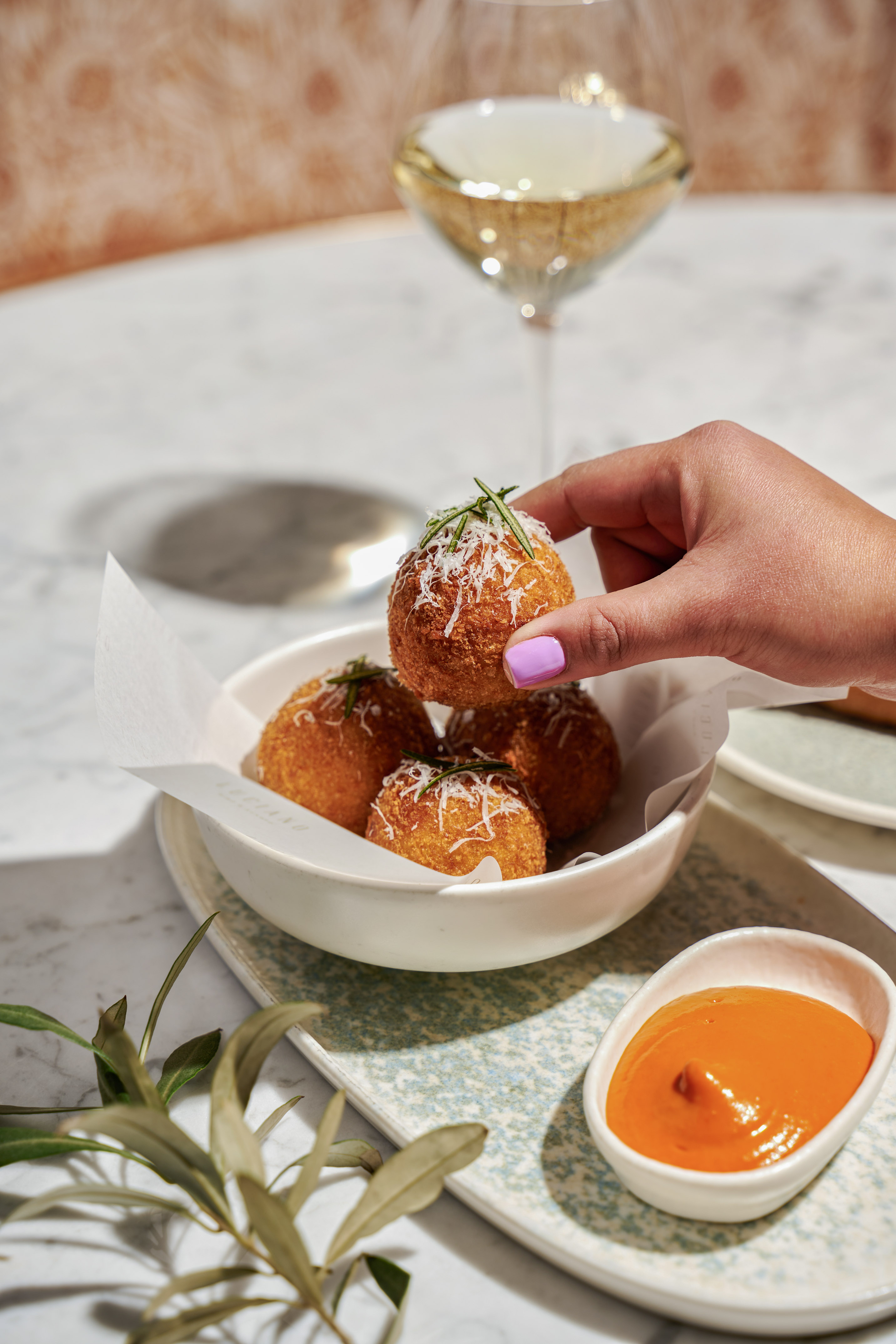 Hand lifting golden fried arancini with parmesan and rosemary from bowl, wine, sauce.