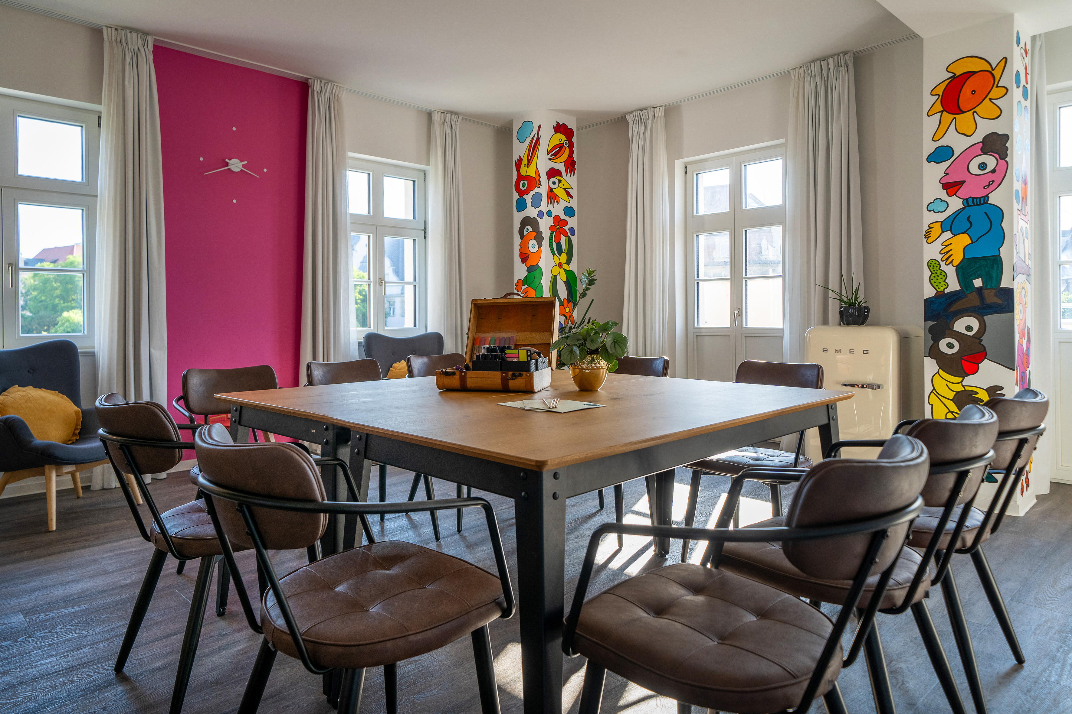 Meeting room with square table, leather chairs, pink wall, and whimsical murals.