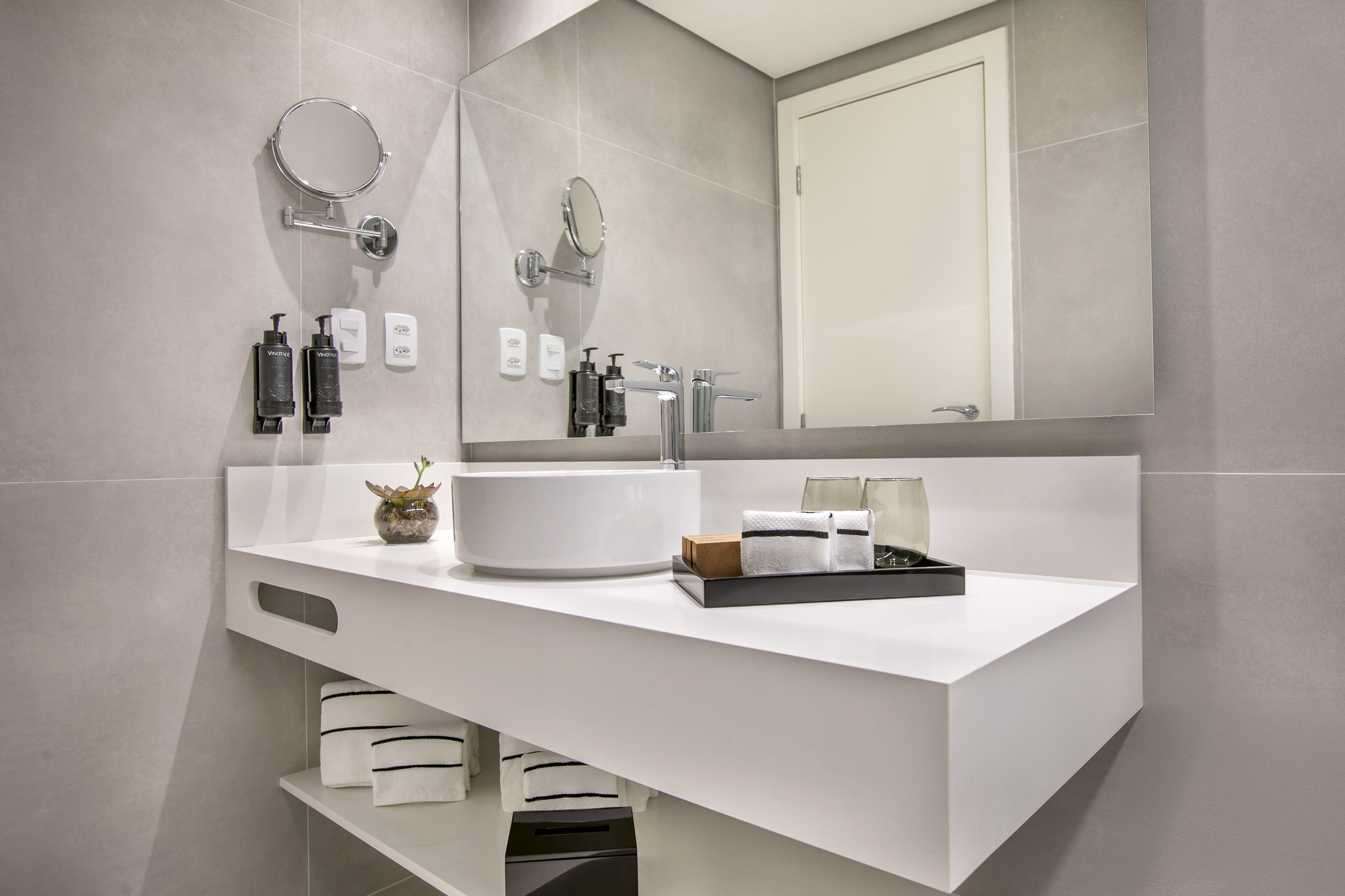 Modern hotel bathroom vanity with white vessel sink, mirror, and grey tiles.