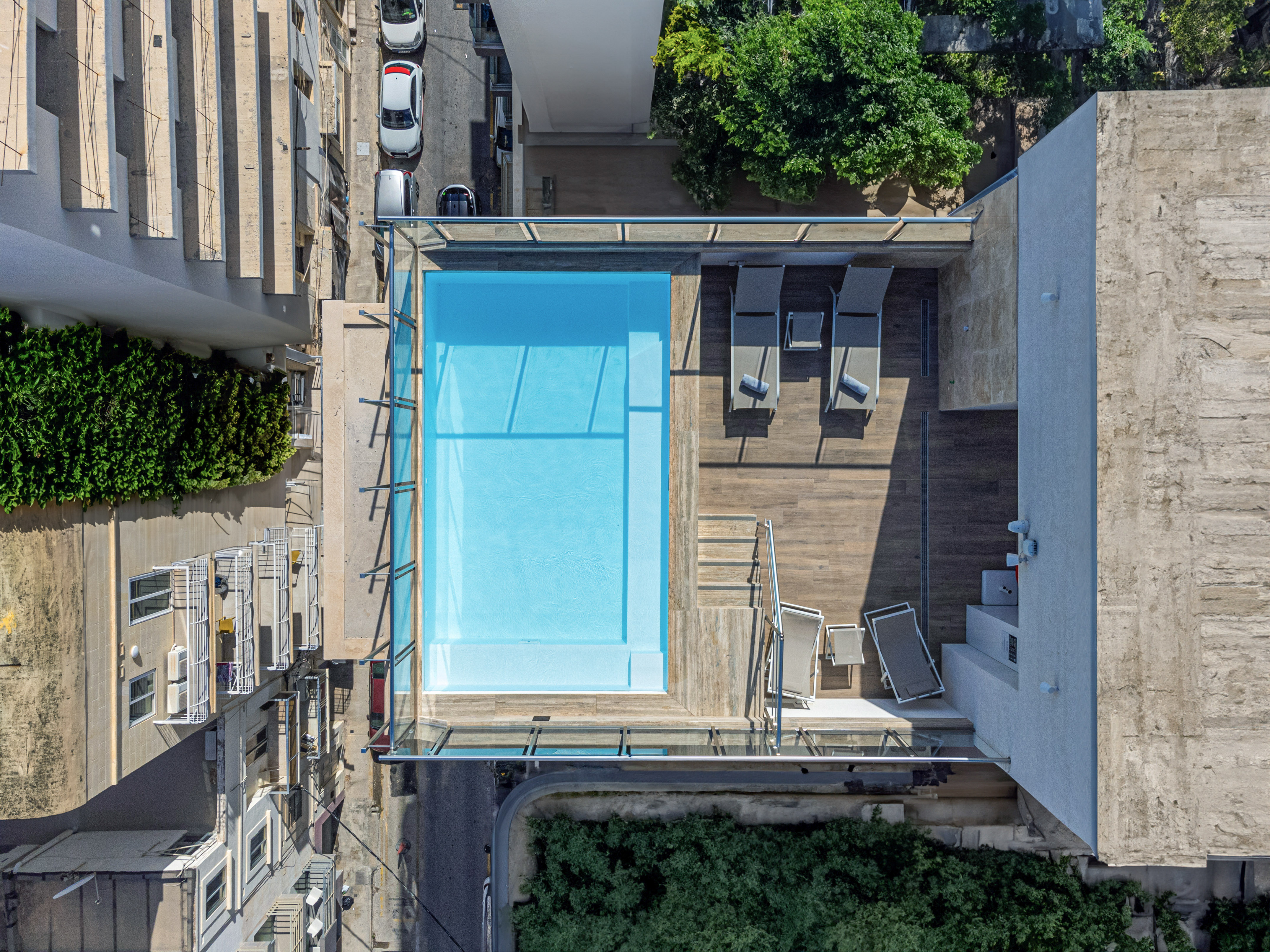 Top-down rooftop swimming pool, wooden deck with sun loungers, urban city.