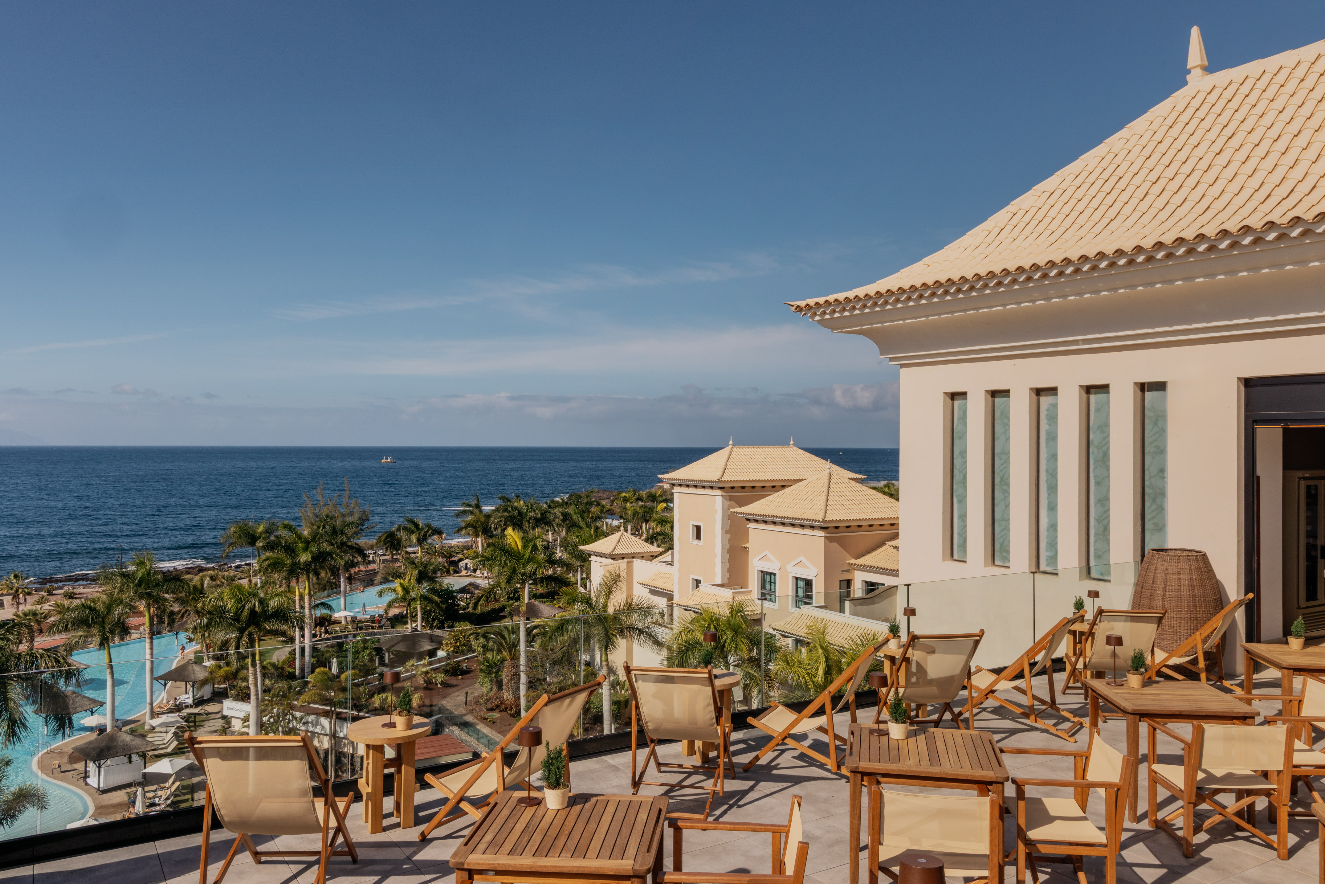 Hotel rooftop terrace overlooking ocean, resort pools, and palm trees.